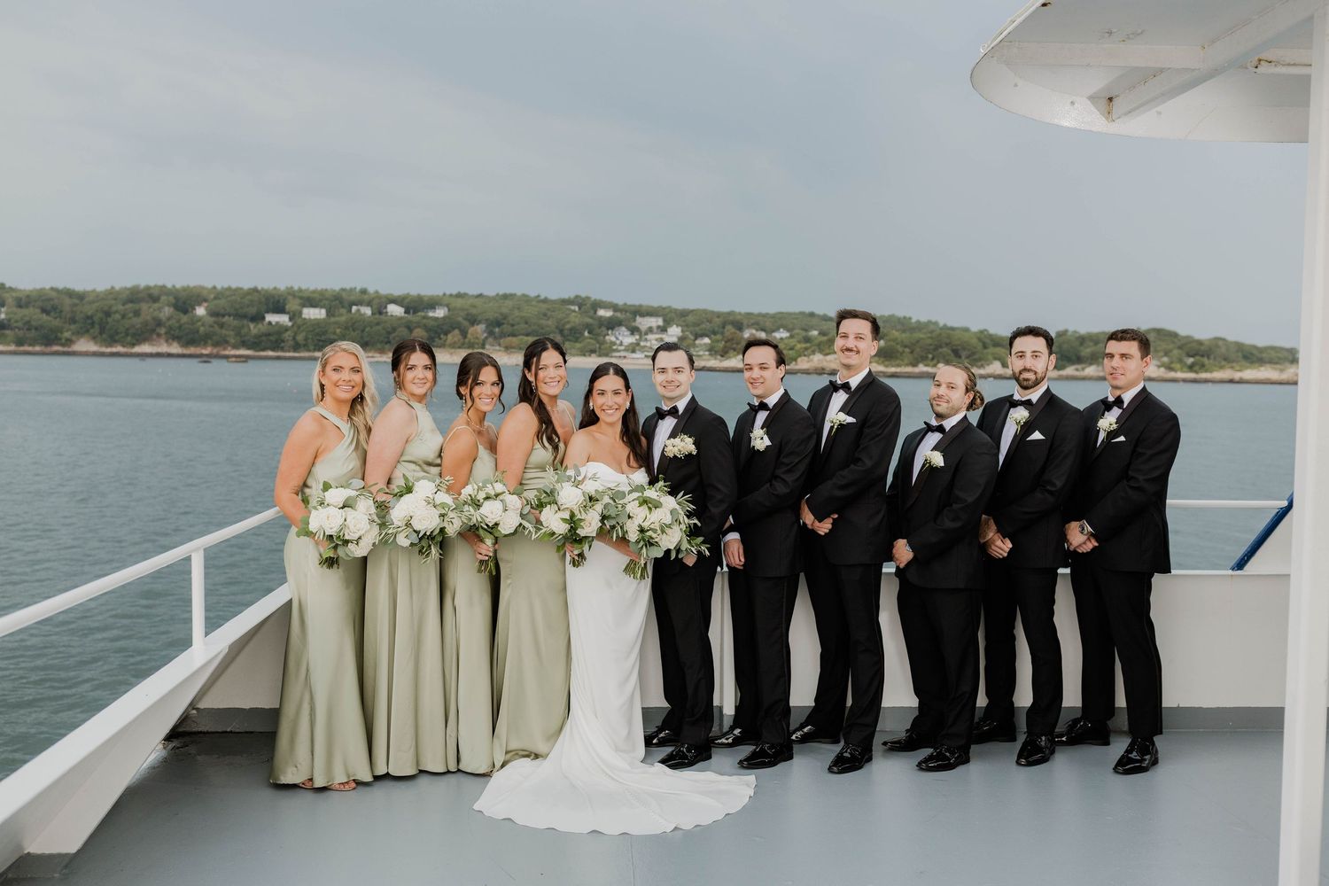 Wedding party in sage bridesmaid gowns and black tuxedos pose together on boat deck by water.