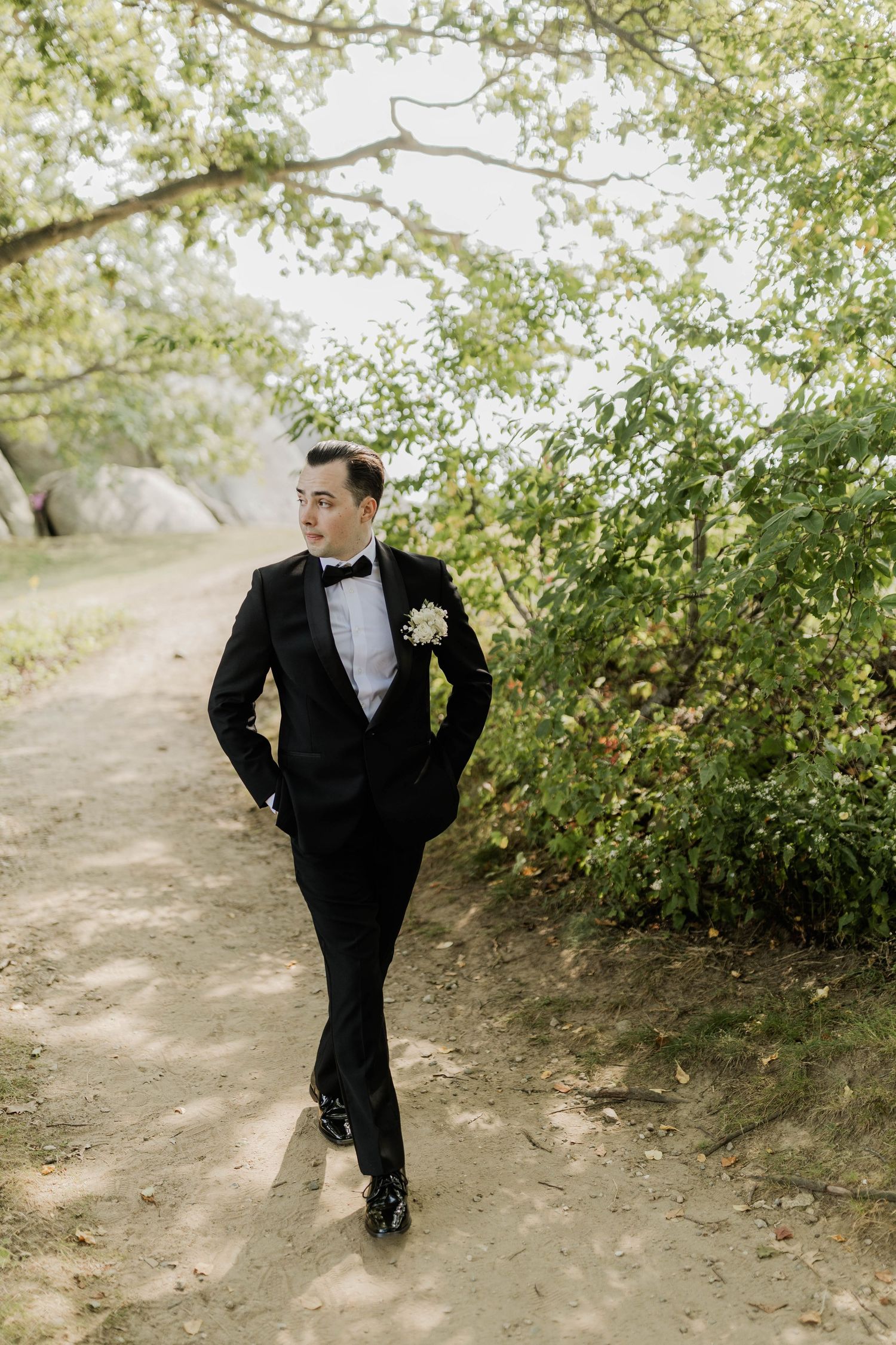 Dapper groom in black tuxedo with boutonniere walks along scenic outdoor path lined with green trees.