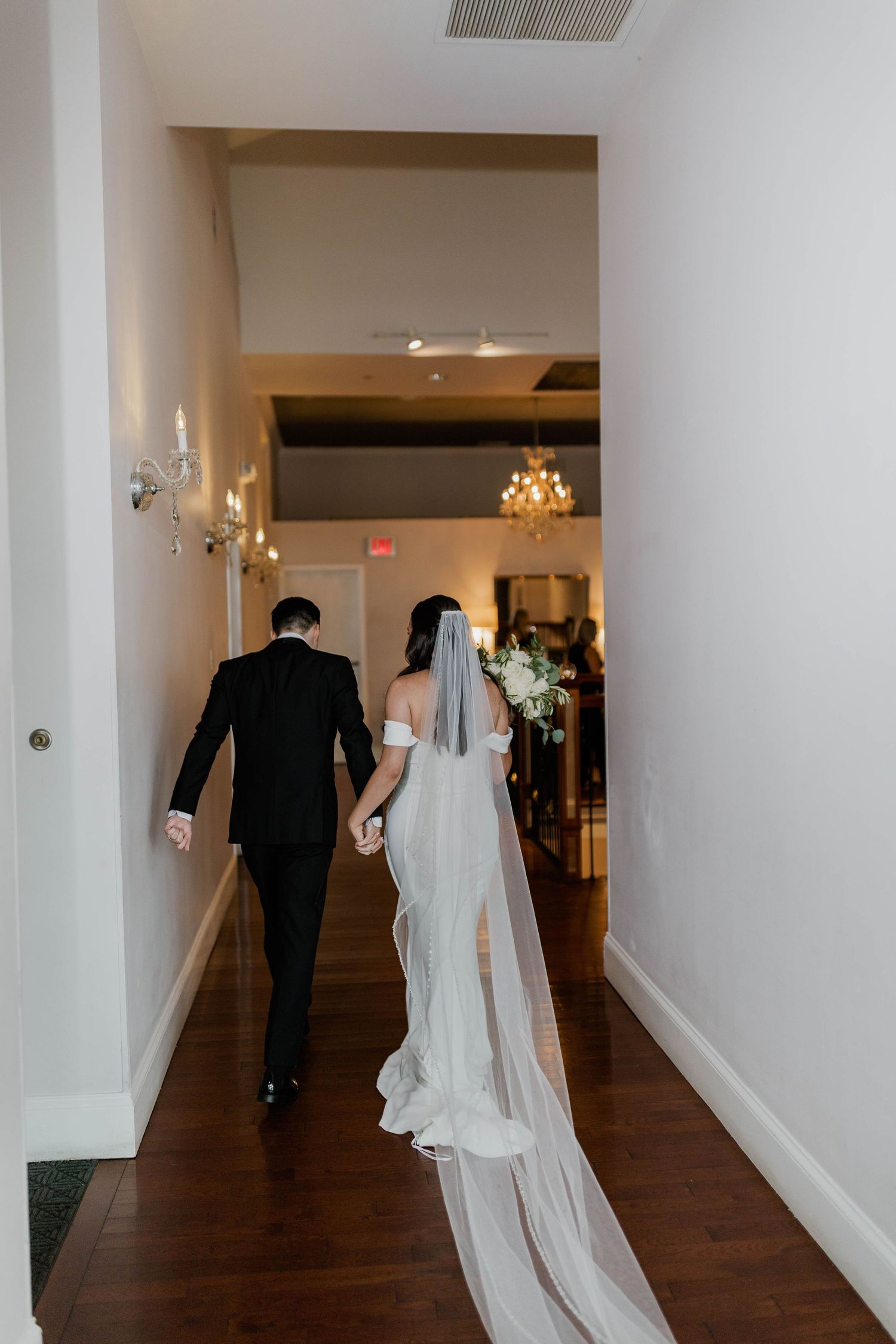 Bride in cathedral veil and groom walk hand-in-hand down hallway toward wedding ceremony.