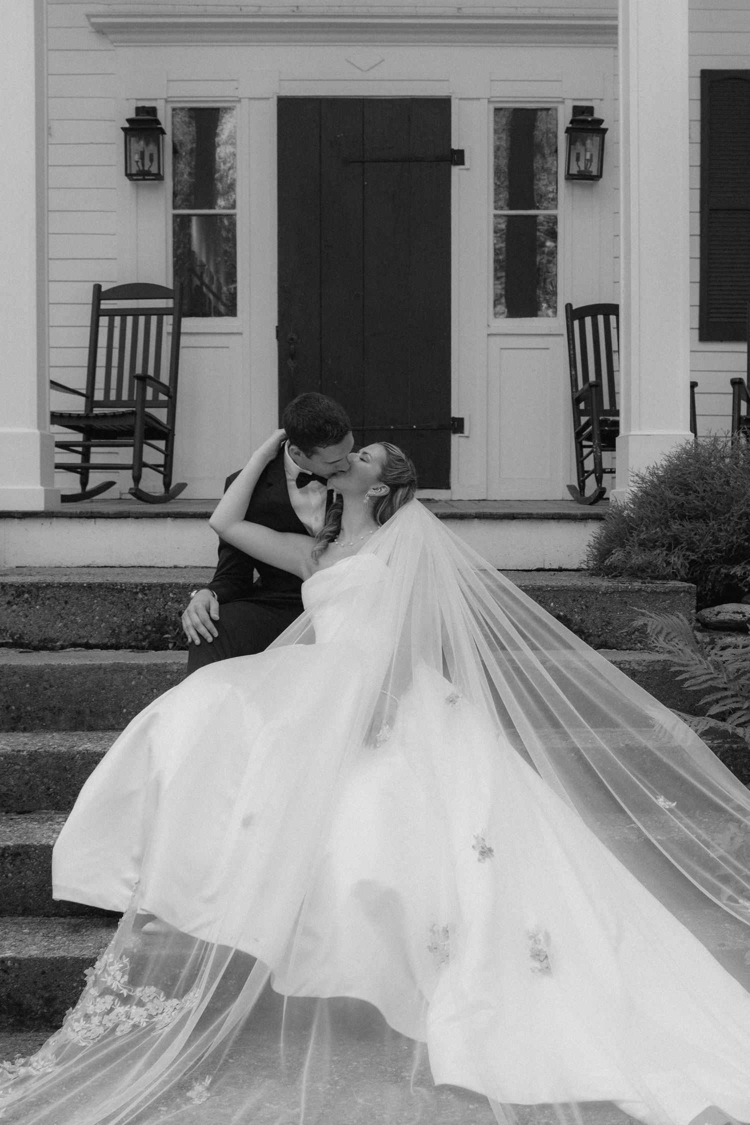 Black and white photo of bride and groom sharing a romantic kiss on steps of white house with rocking chairs.