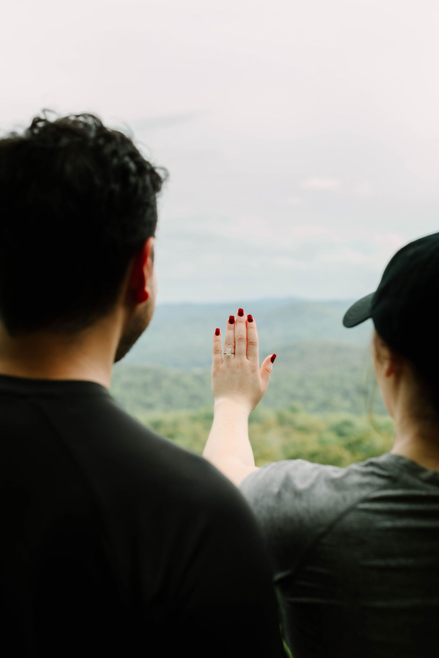 Couple admiring scenic forest view from overlook, woman displaying engagement ring with red nails.