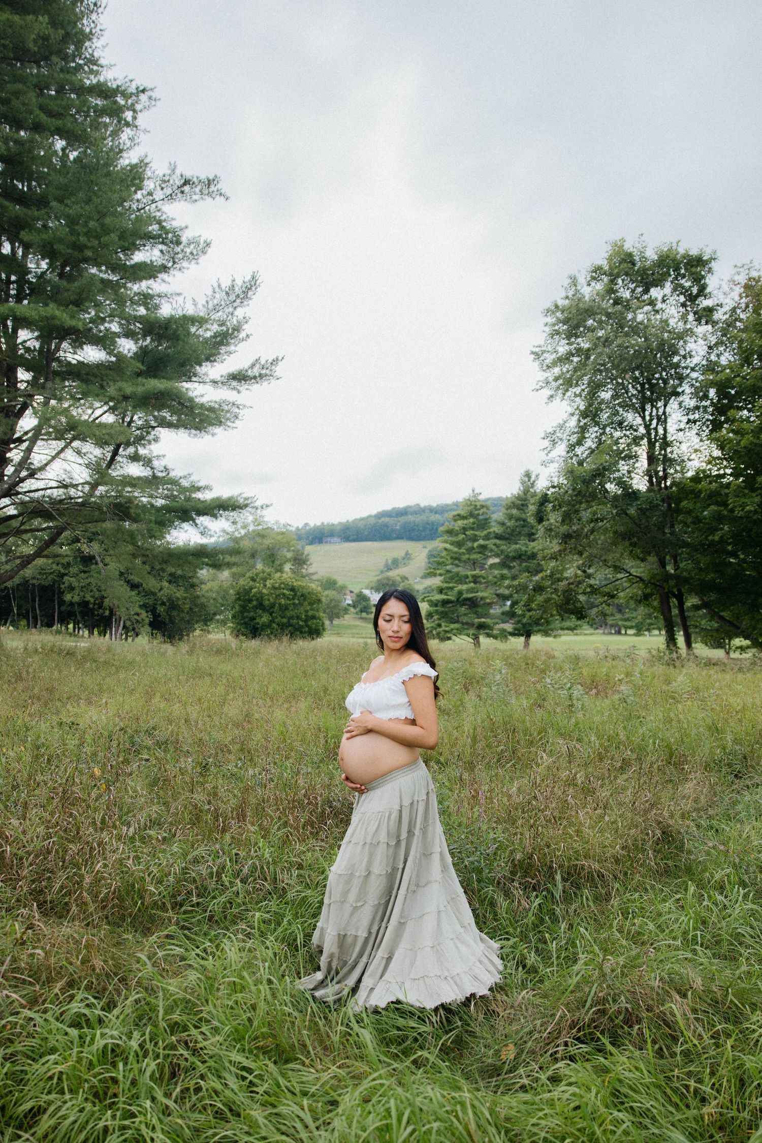 Pregnant woman in white top and gray skirt cradling baby bump in lush green meadow with trees and hills.