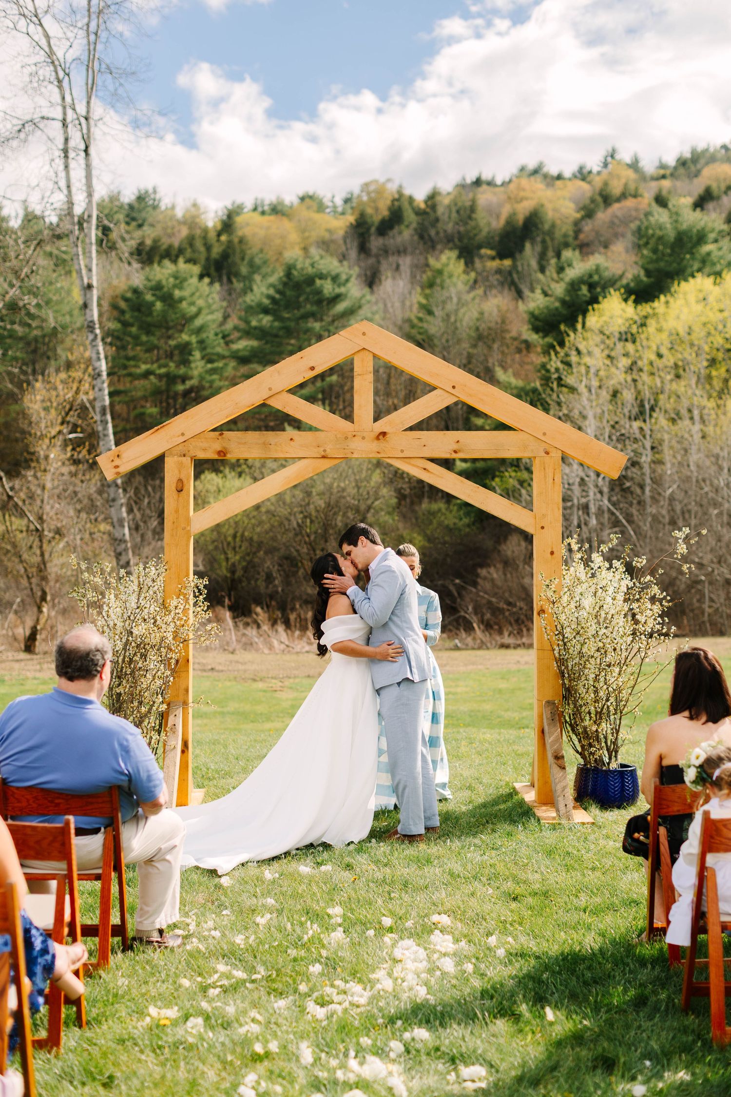 Newlyweds share their first kiss under a rustic wooden arch during an outdoor wedding ceremony with forest backdrop.