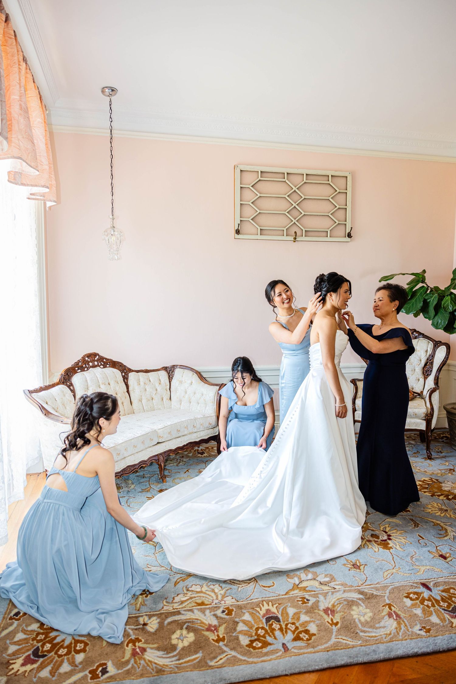 Bridesmaids in blue dresses help a bride in a white gown prepare in an elegant pink room before her wedding.