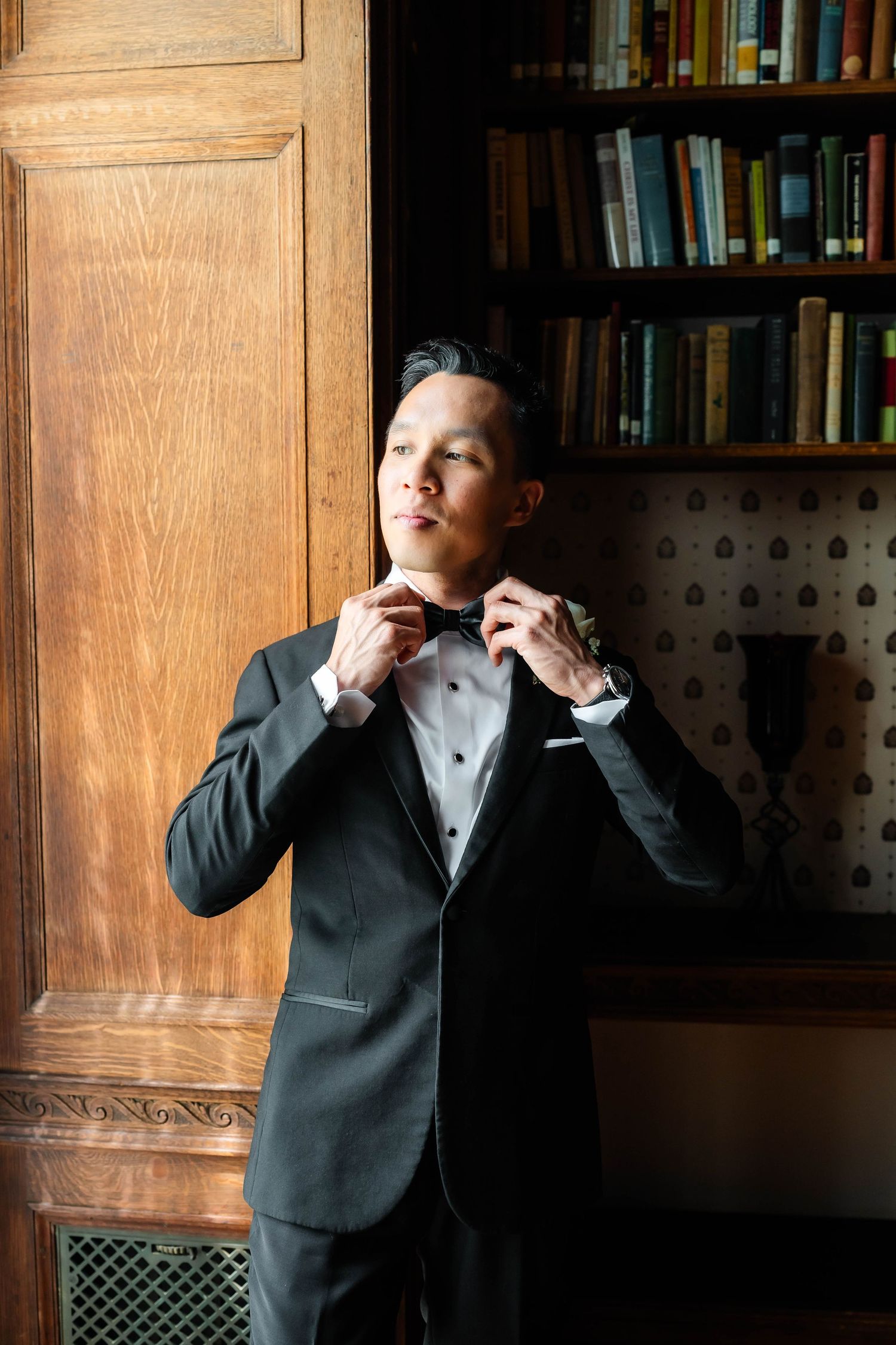 Dapper Asian groom in black tuxedo adjusting bow tie while standing beside wooden bookshelf in library.