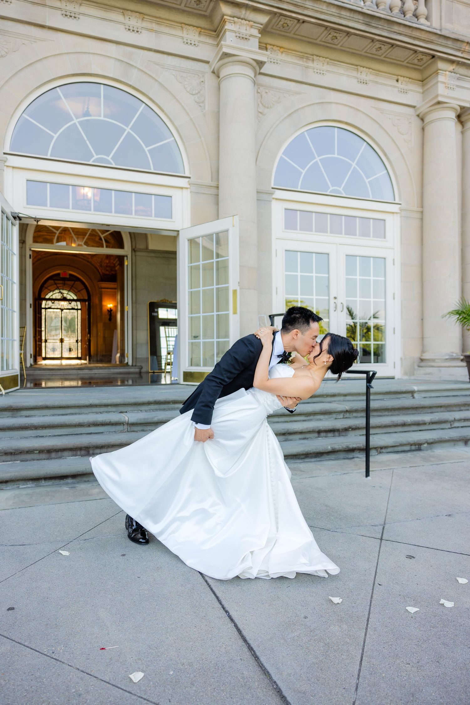 Groom dipping bride in romantic kiss outside grand neoclassical building with arched windows.