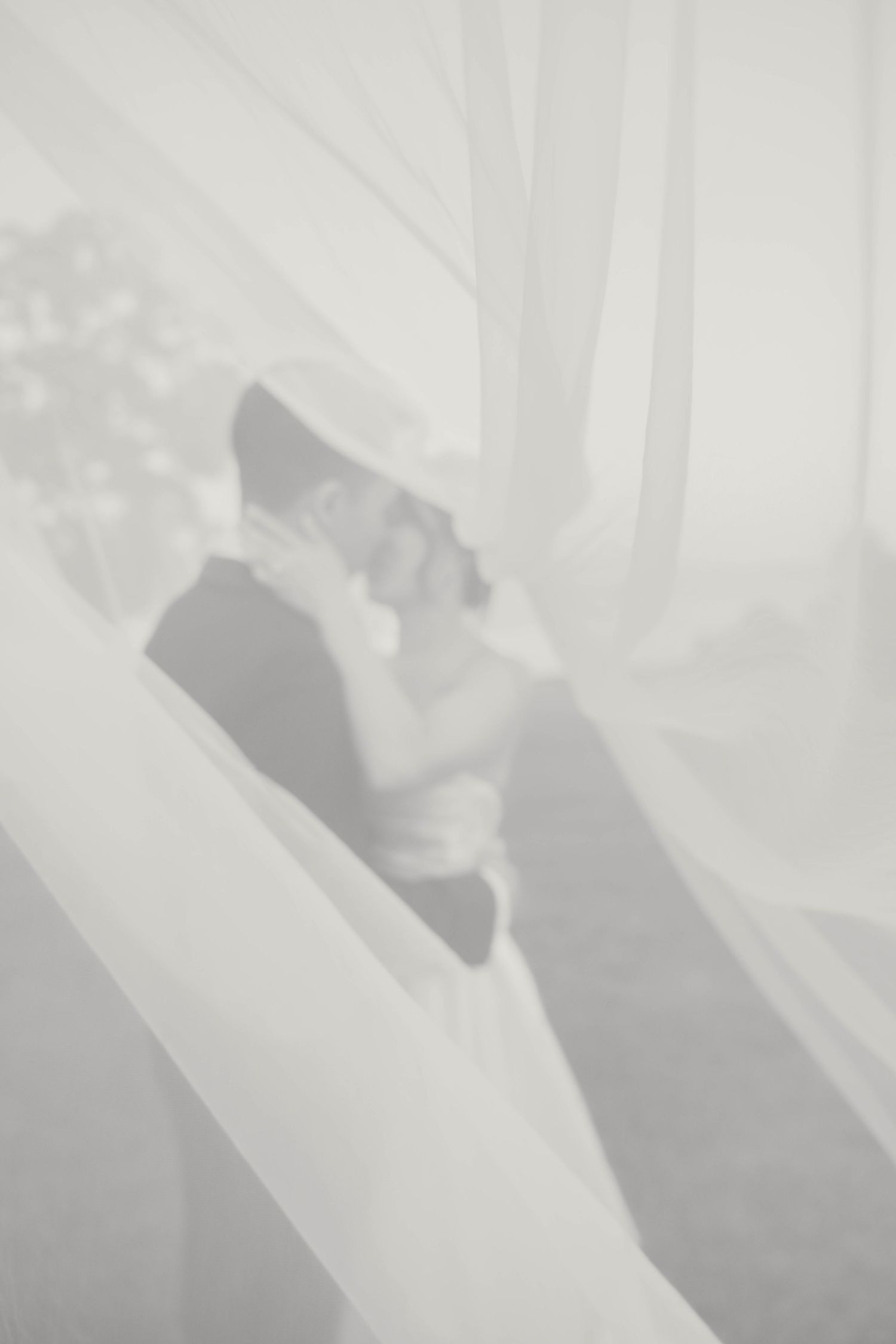 Dreamy silhouette of bride and groom kissing through flowing white sheer fabric.