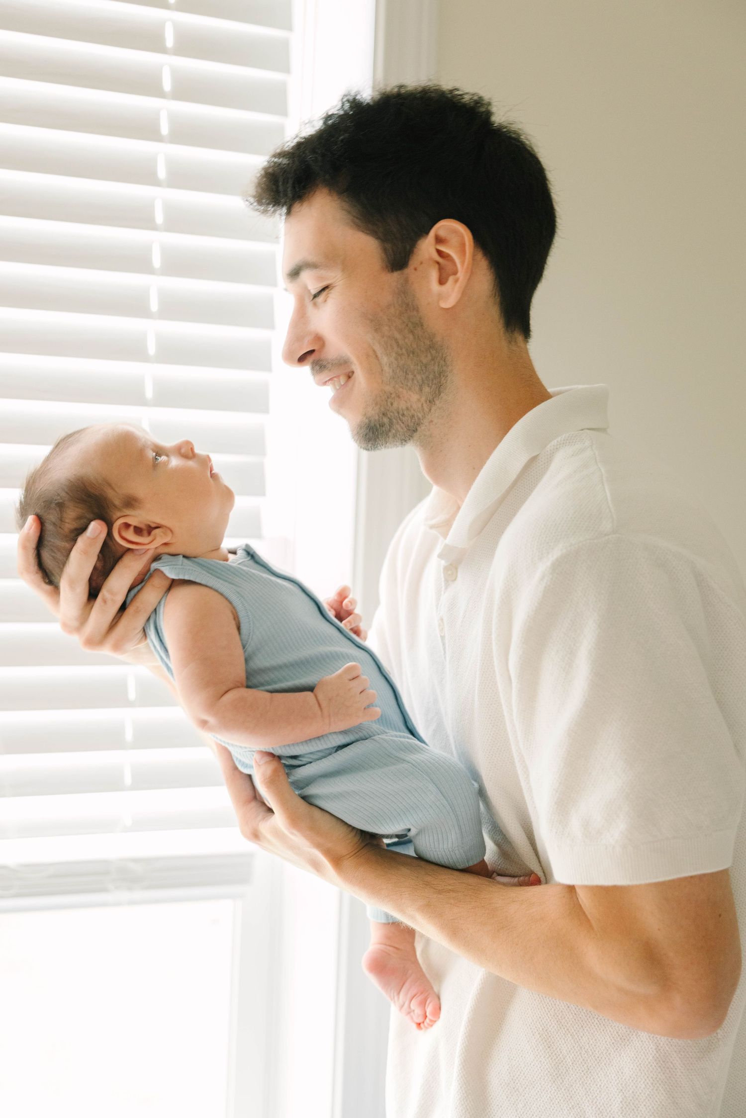 Smiling father in white shirt holding newborn baby in blue swaddle near a bright window.