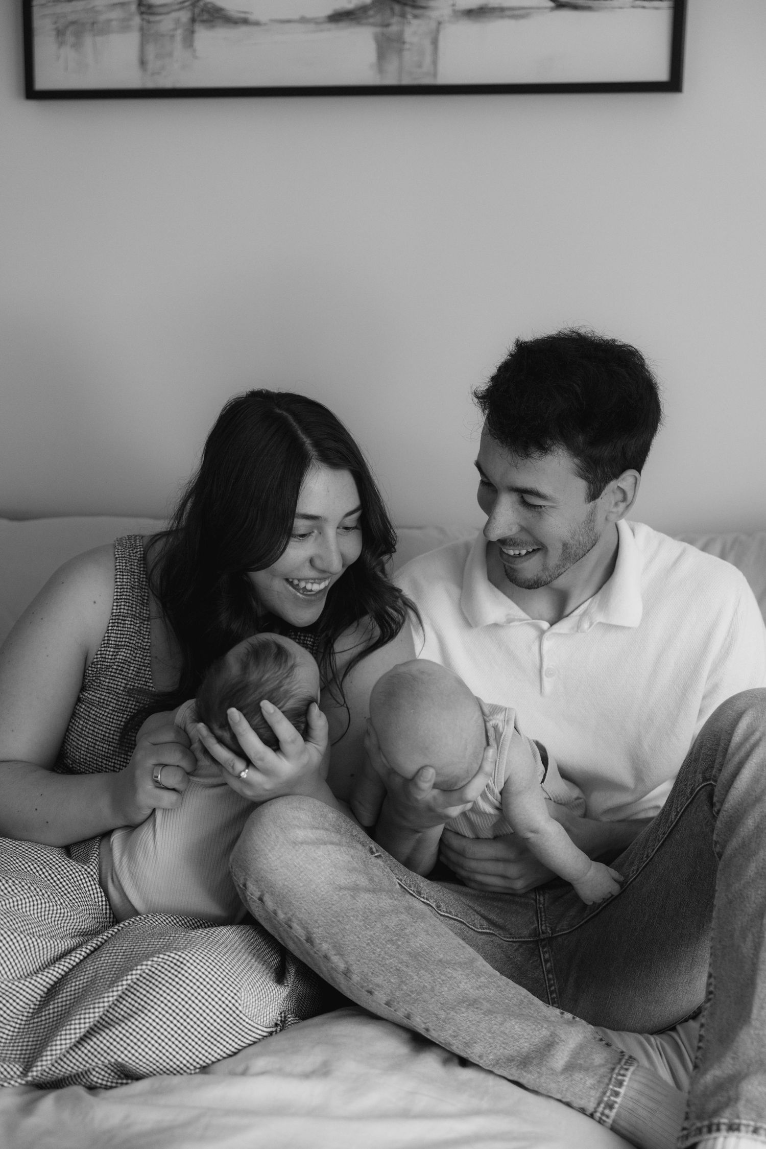Black and white photo of smiling parents sitting on a bed together holding their newborn baby.