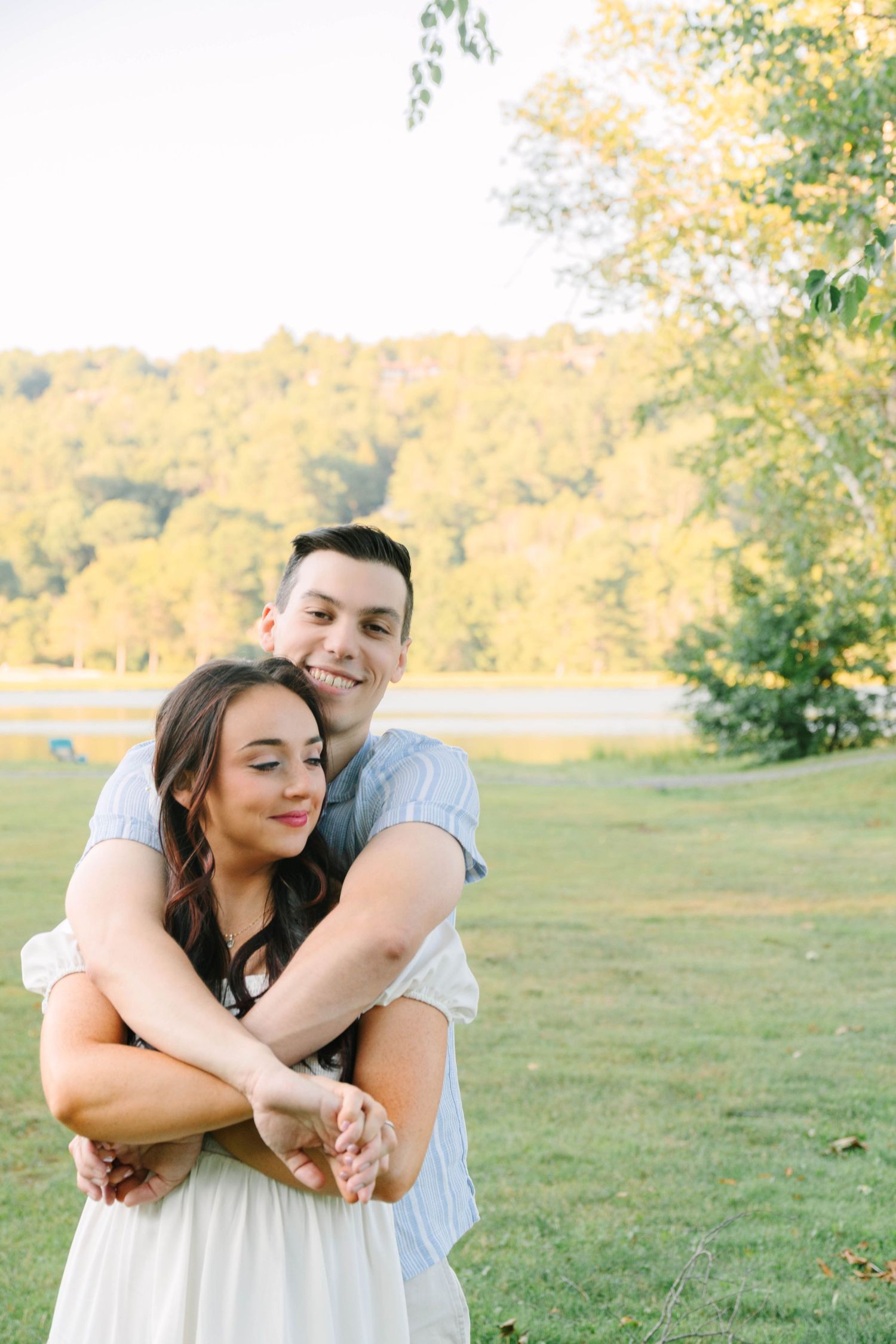 A couple embraces outdoors near a lake, man hugging woman from behind, both smiling, surrounded by lush green trees.