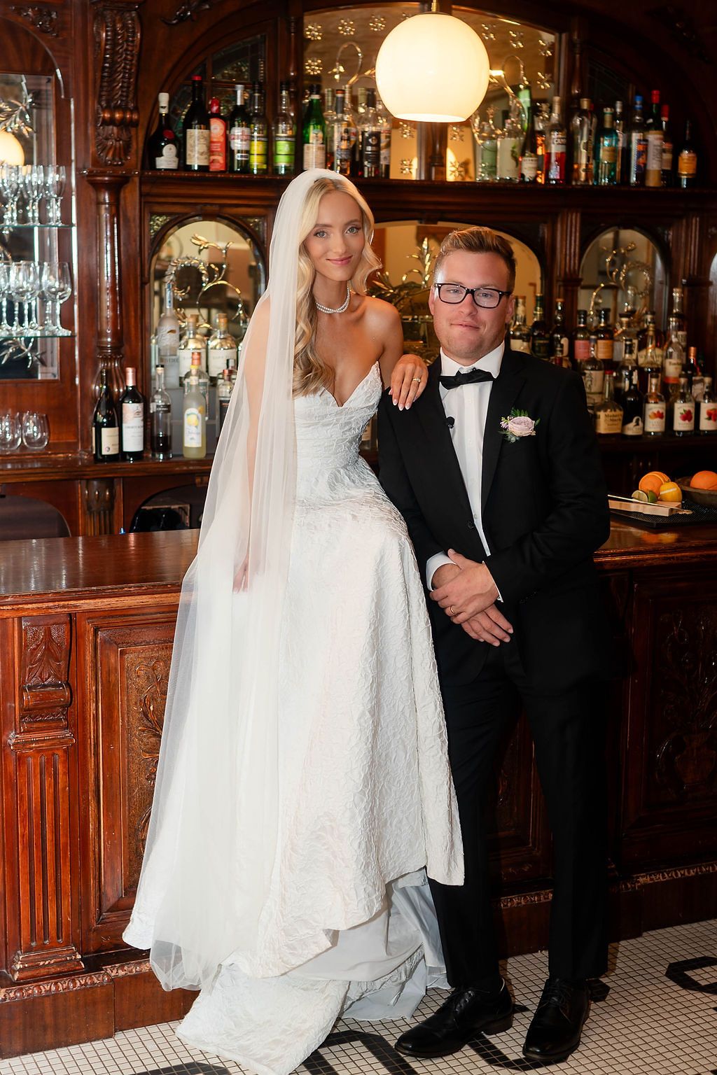 Bride in white lace gown with veil poses with groom in black tuxedo at an elegant wooden bar.