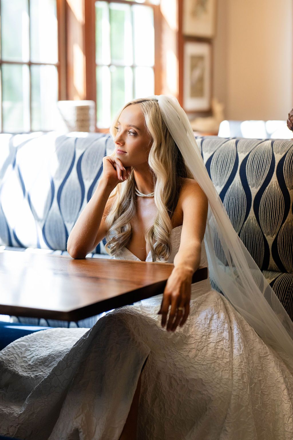 A bride in a white gown and veil sits thoughtfully at a wooden table on a blue patterned sofa near a sunlit window.
