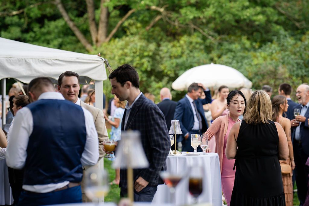 Wedding guests mingle outdoors under white tents with drinks during a sunny garden cocktail hour.