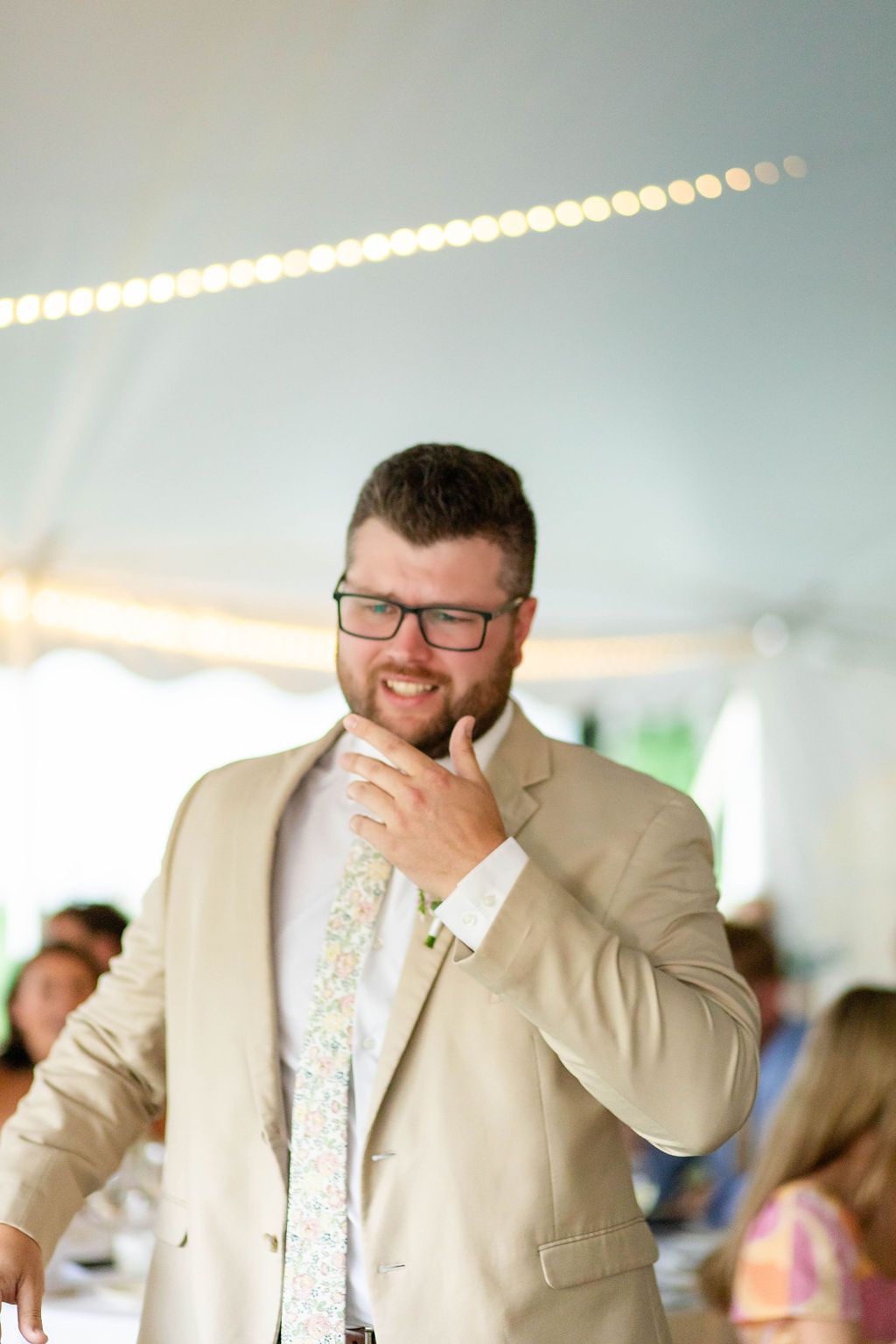 Smiling male wedding guest in tan blazer and floral tie enjoys the reception under a white tent.