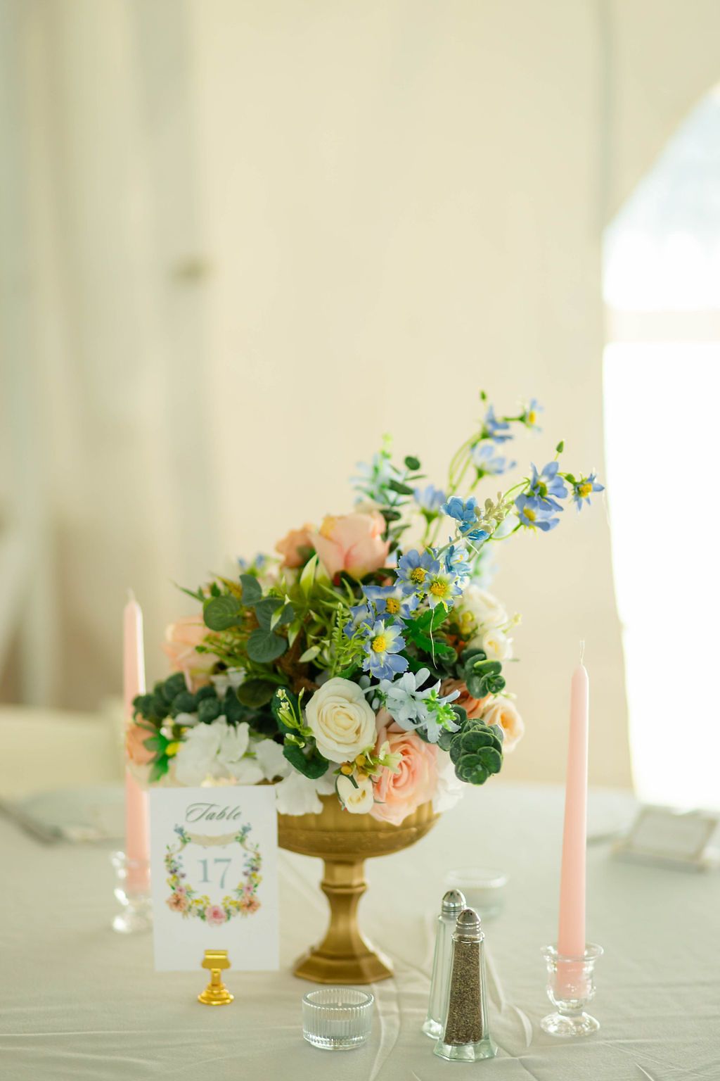 Elegant wedding centerpiece with pink and blue flowers in gold urn flanked by pink candles.