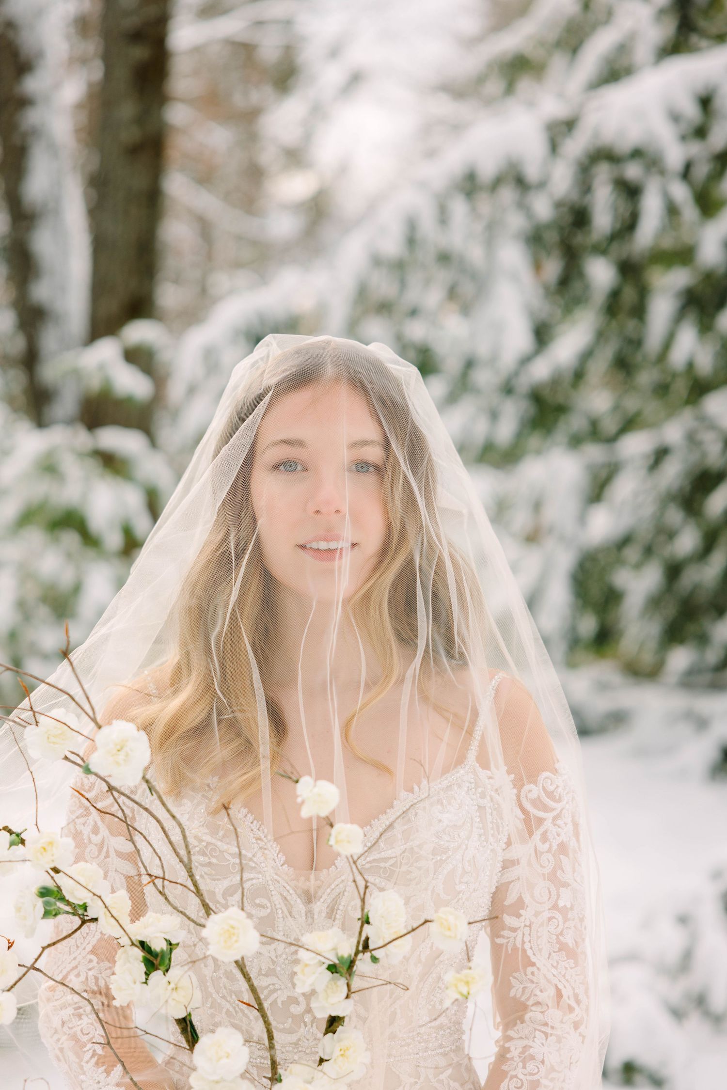 Bride in lace gown with veil over face holding white floral bouquet in snowy forest.