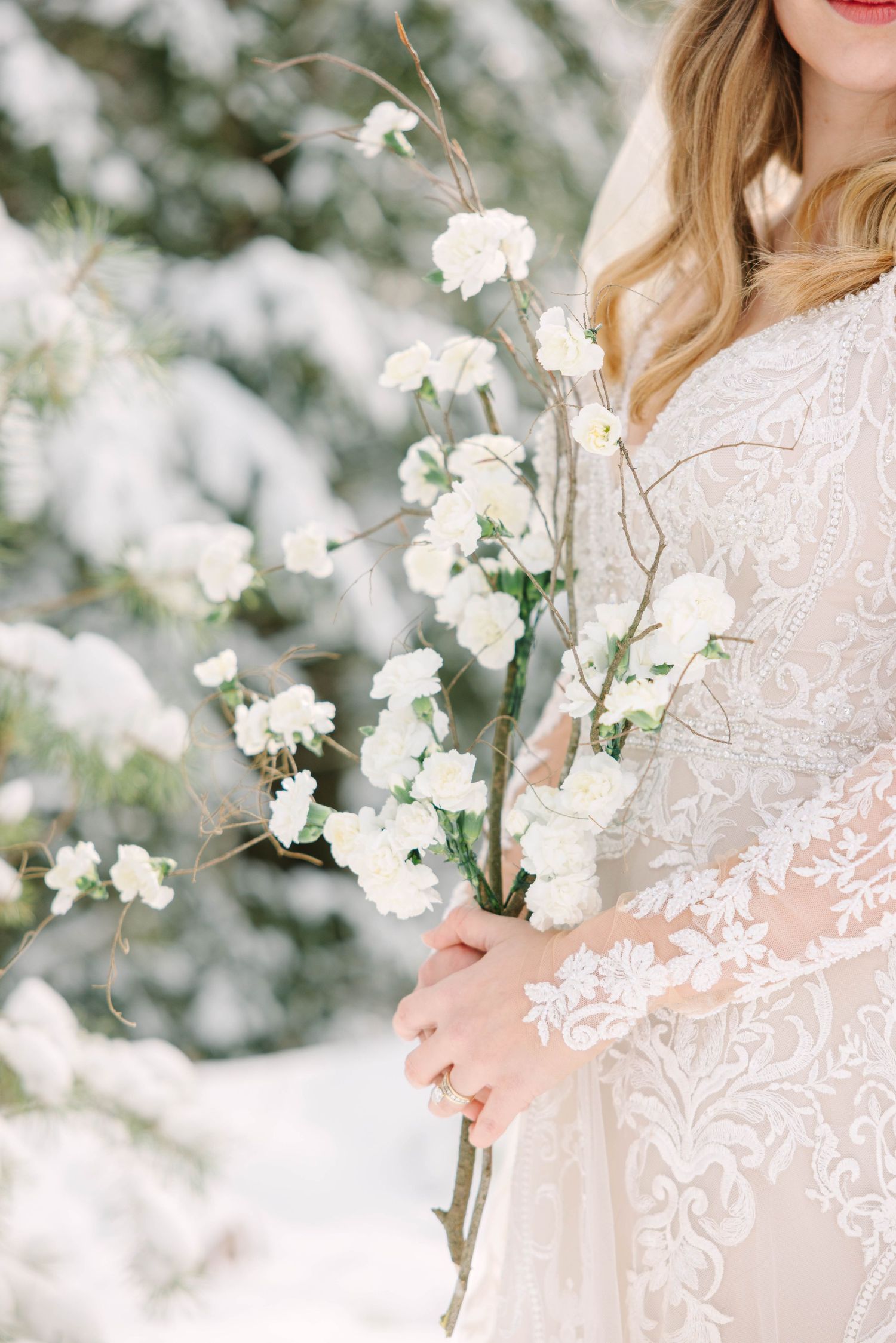 Bride in lace gown holding white floral branches in snowy winter forest setting.