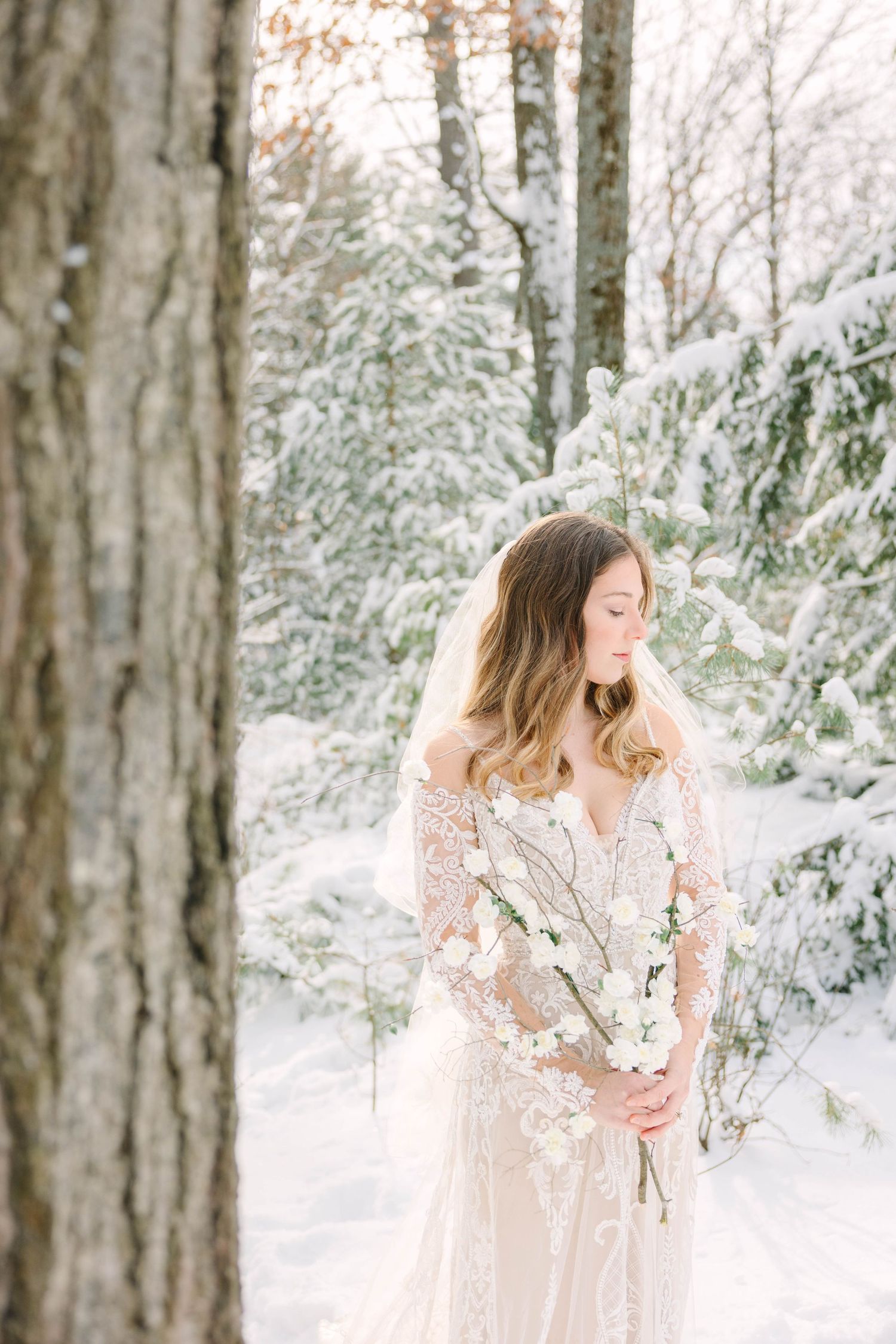 Bride in lace off-shoulder gown holding white floral bouquet in a serene snow-covered winter forest.