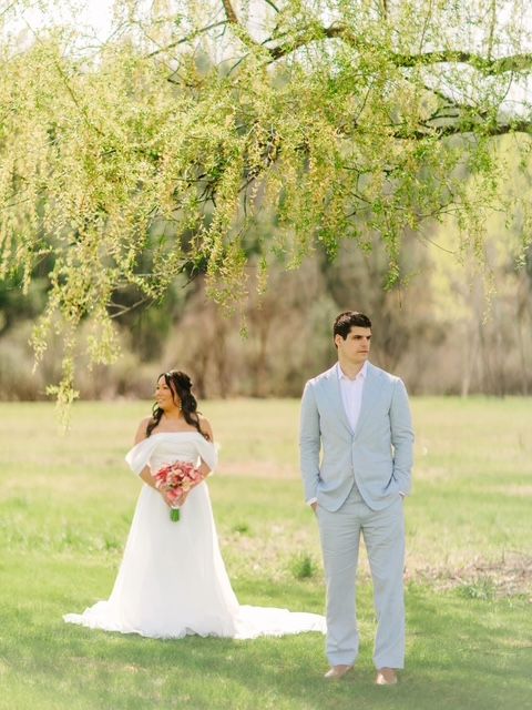 Bride in off-shoulder white gown holding pink bouquet stands near groom in light blue suit under weeping willow tree.