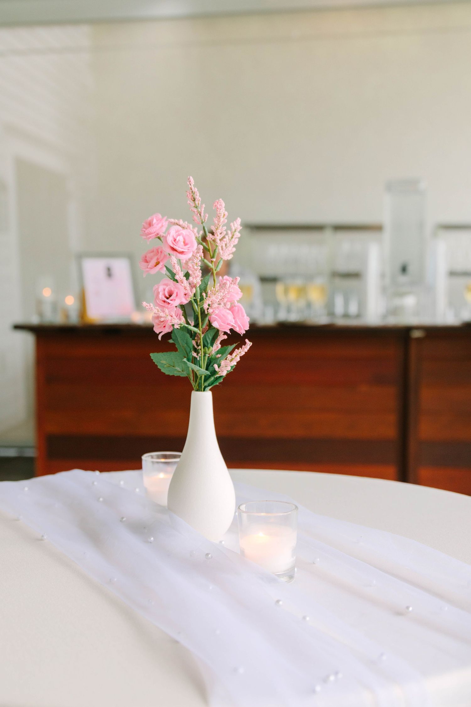 White bud vase with pink roses on white table runner with pearl accents and votive candles at reception.