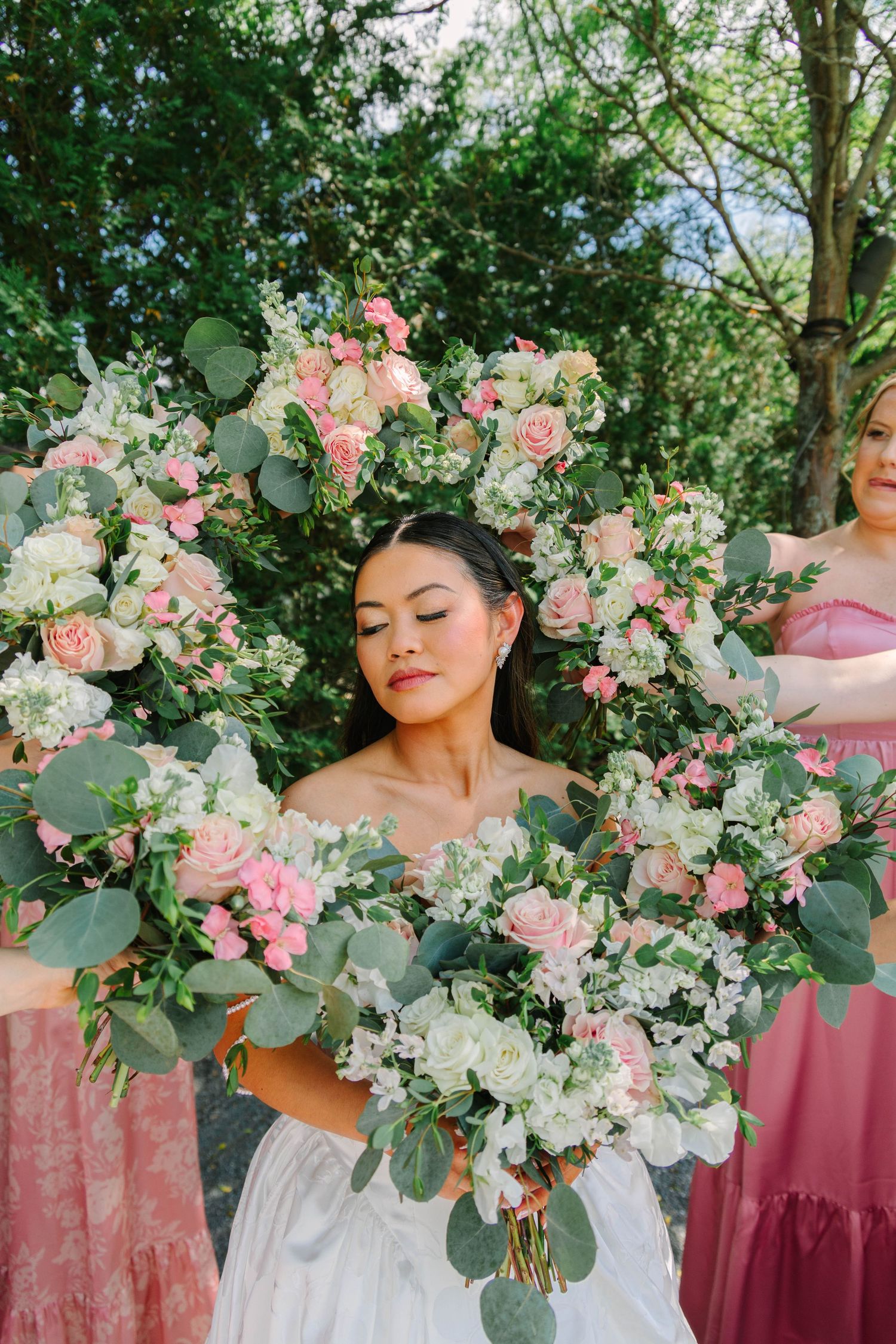 Bride framed by bridesmaids holding pink and white rose bouquets forming a floral arch outdoors.