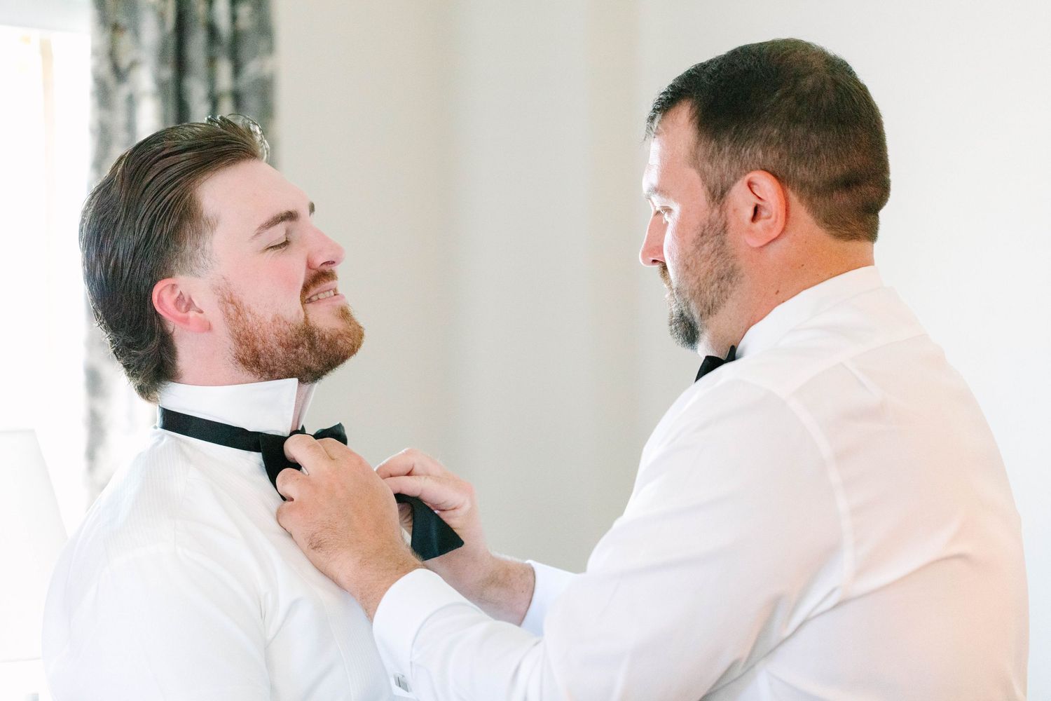 Two men in white dress shirts as one carefully ties the other's black bow tie before the wedding.