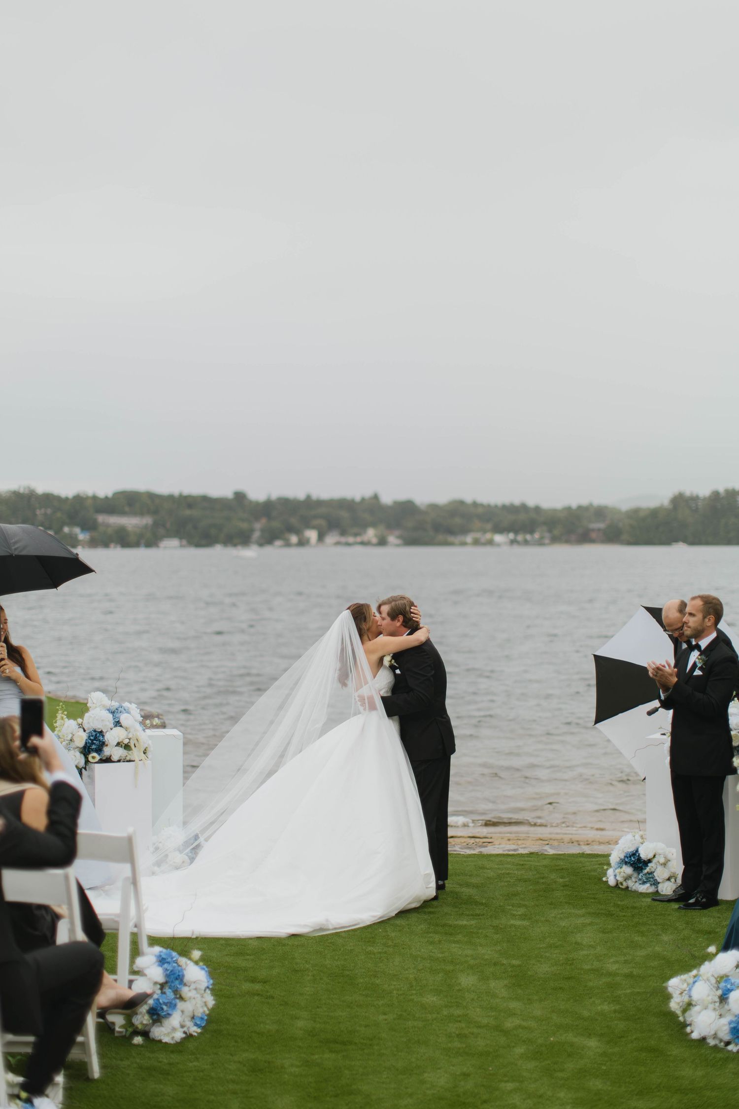 Bride and groom share first kiss at outdoor lakeside wedding ceremony on rainy day with guests holding umbrellas.