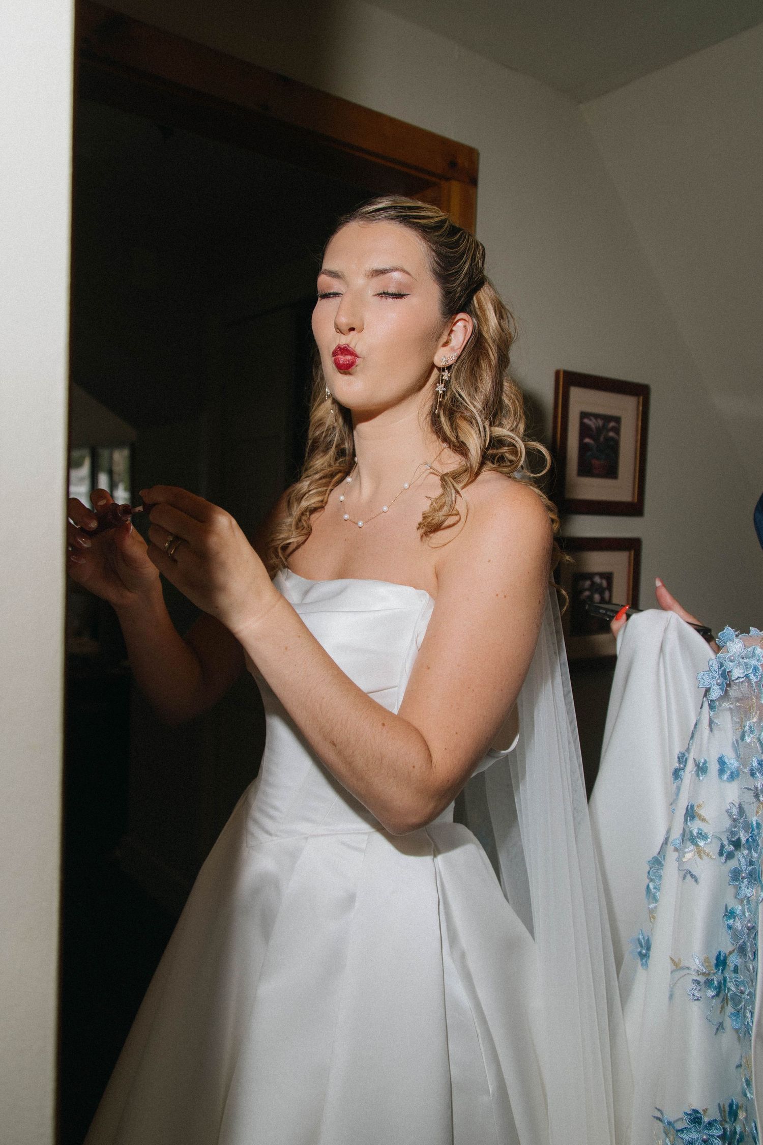 Bride in white strapless gown blowing a kiss while getting ready, with floral dress visible nearby.