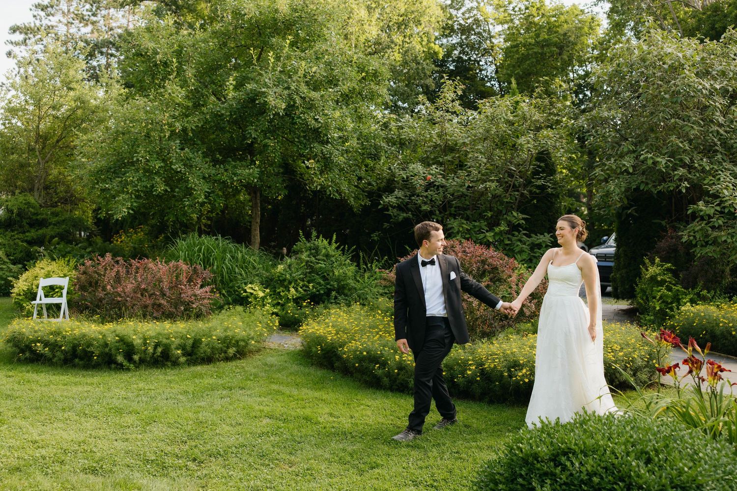 Bride and groom walk hand in hand through a lush green garden on their wedding day.