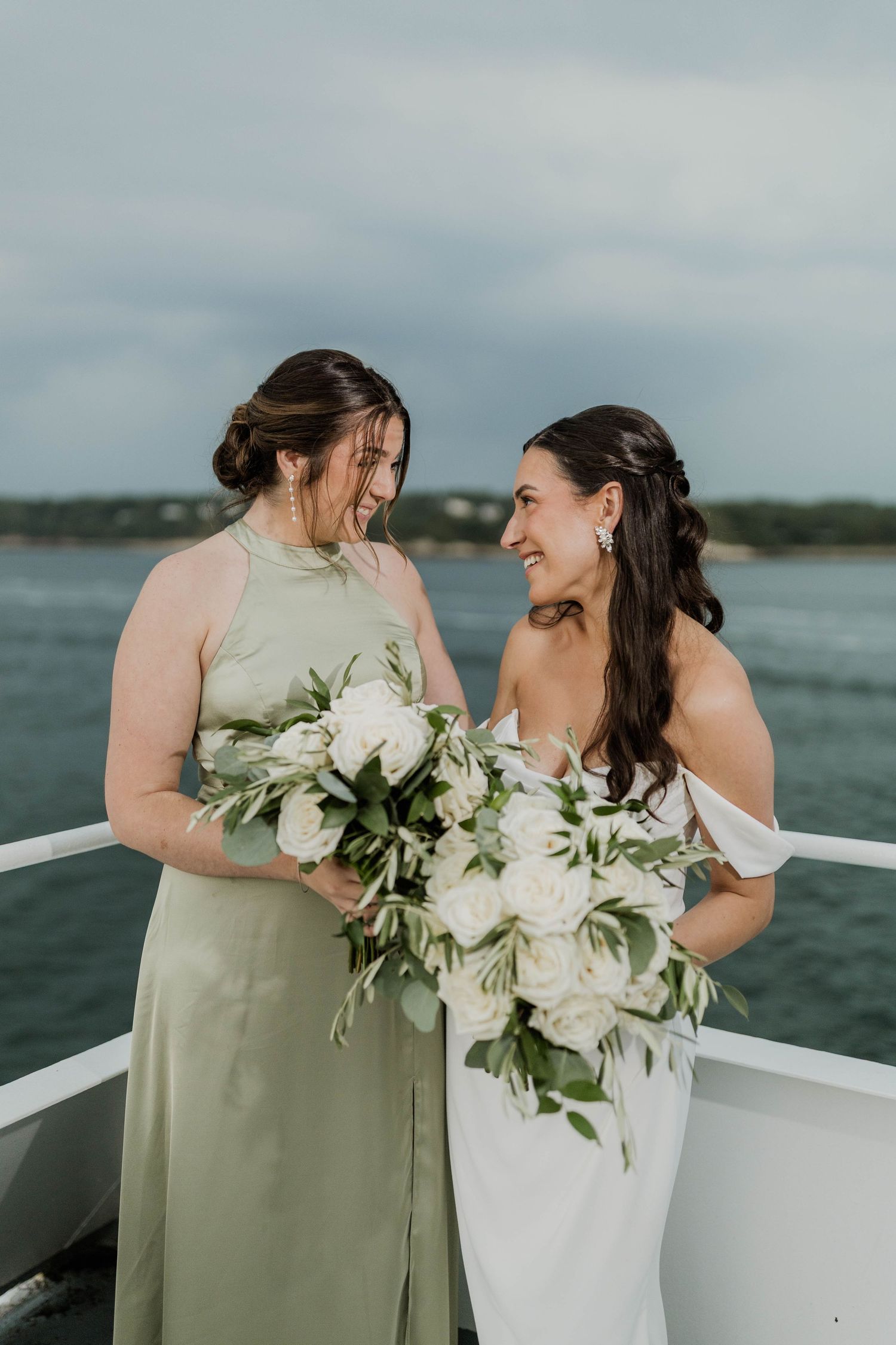 Bride in white gown and bridesmaid in sage green dress laugh together on a boat holding white floral bouquets.