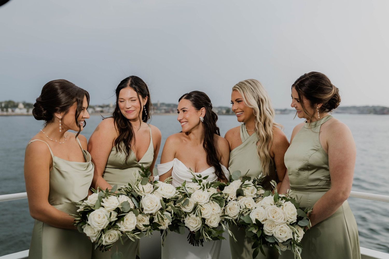 Bride and four bridesmaids in sage green dresses laugh together holding white rose bouquets on a boat.
