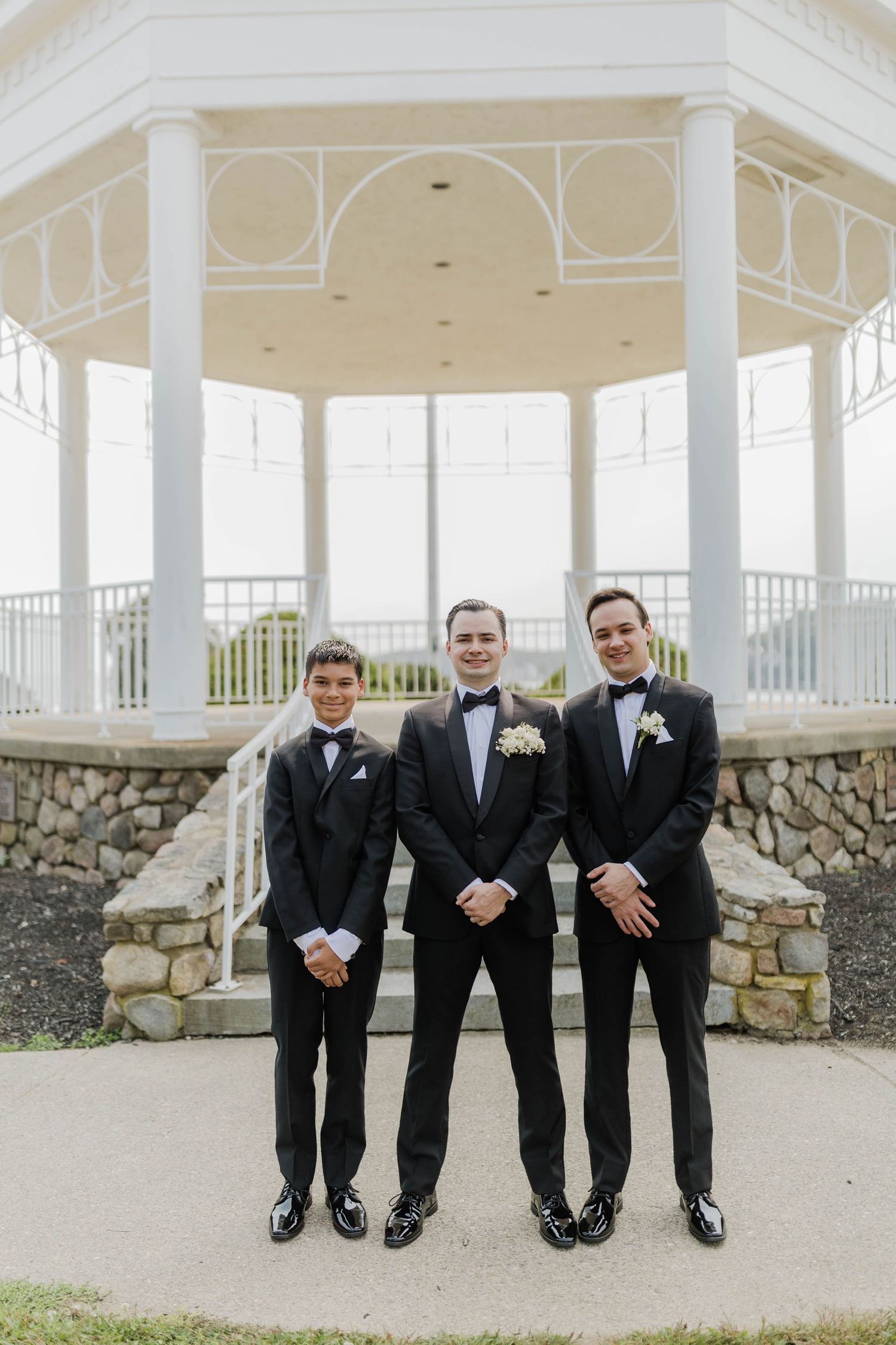 Three groomsmen in black tuxedos with boutonnieres pose on steps before a white gazebo at an outdoor wedding venue.
