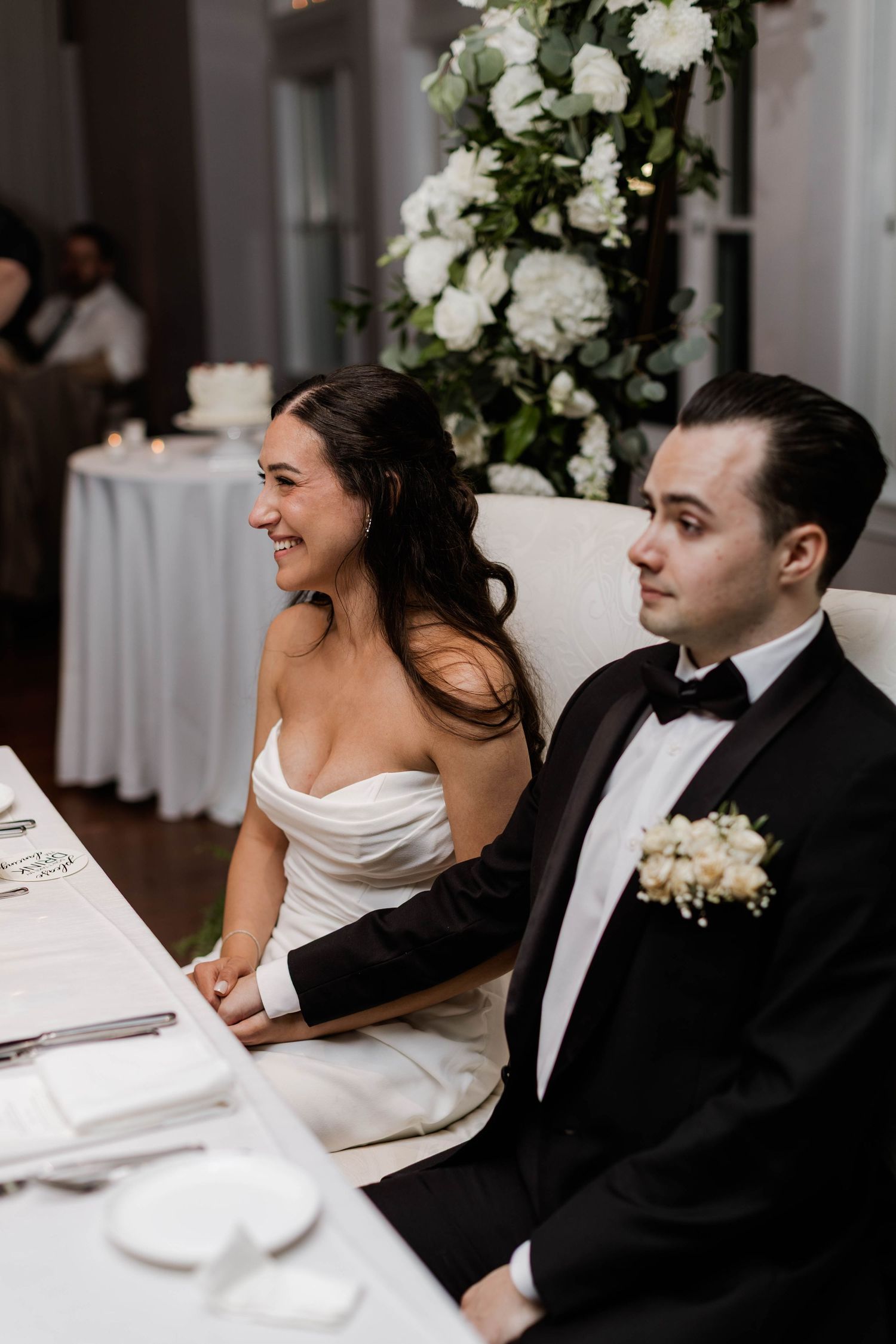 Smiling bride in strapless white gown and groom in black tuxedo seated together at wedding reception.