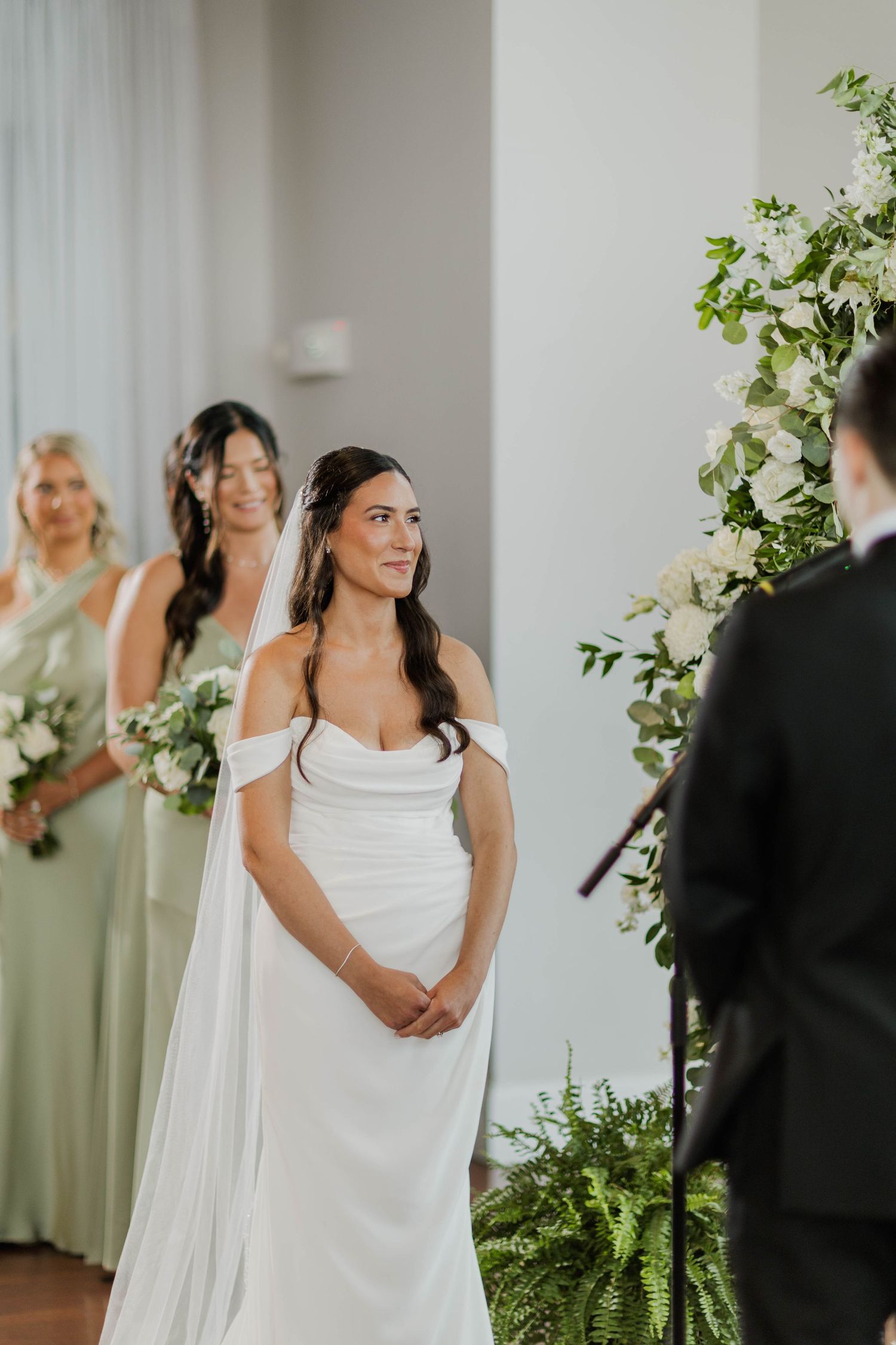 Bride in white off-shoulder gown with veil stands at altar with sage green bridesmaids during ceremony.