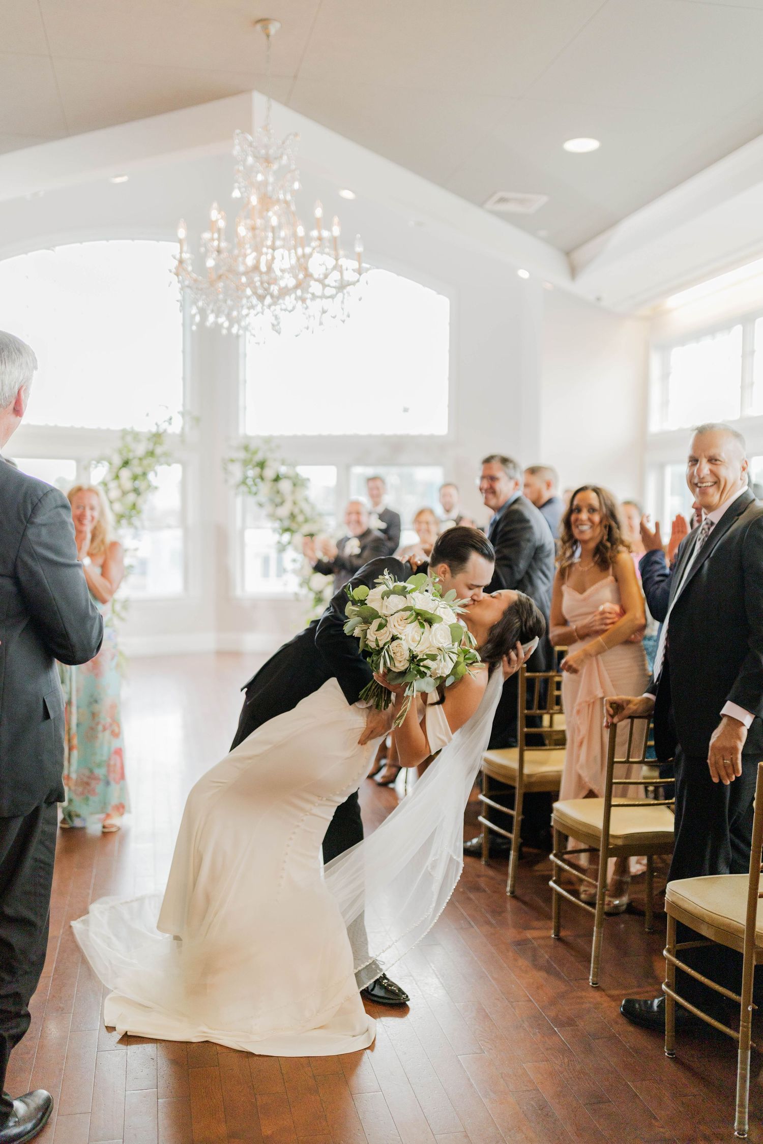 Groom dips bride for a kiss in the aisle after wedding ceremony as guests cheer in elegant venue.