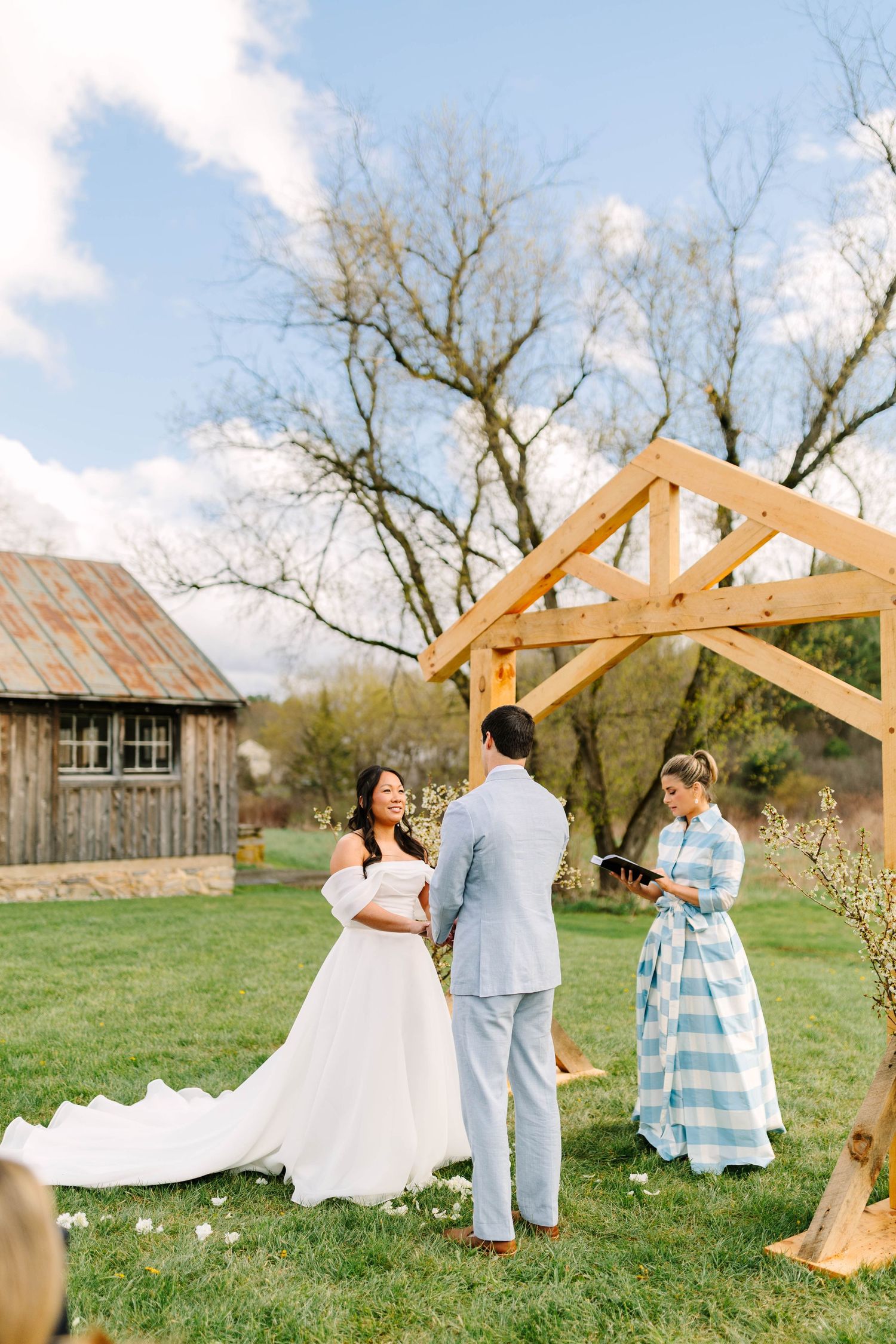 Bride and groom exchanging vows outdoors under wooden arch structure with officiant in blue striped dress.