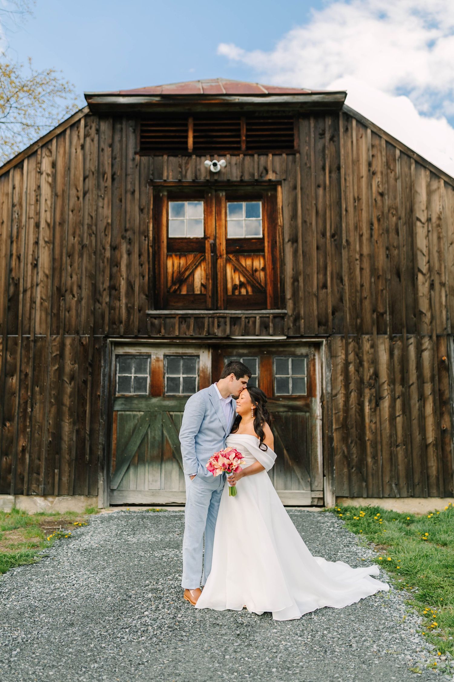 Bride and groom sharing romantic kiss in front of rustic dark wood barn with pink calla lily bouquet.