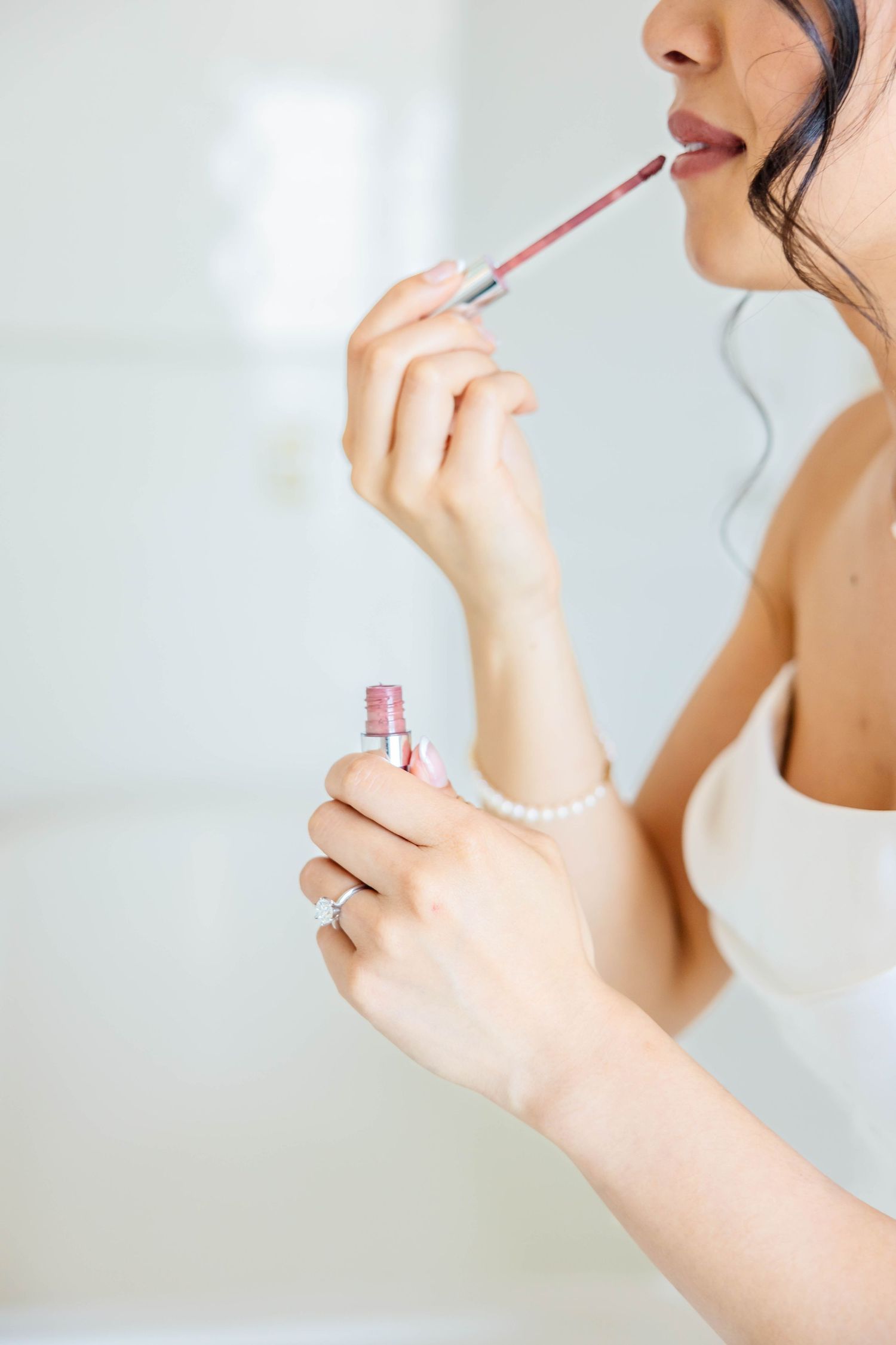 Bride applying pink lip gloss while wearing a pearl bracelet and diamond engagement ring on her wedding day.