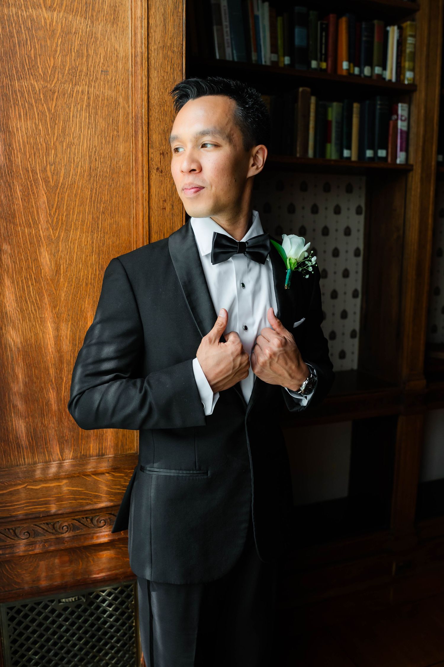 Dapper groom in black tuxedo with boutonniere adjusting lapels in front of wooden bookshelf.