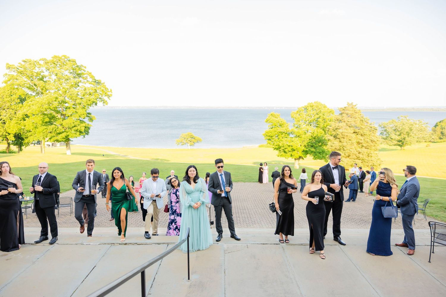 Wedding guests mingling on terrace steps with scenic waterfront lawn and clear sky in background.
