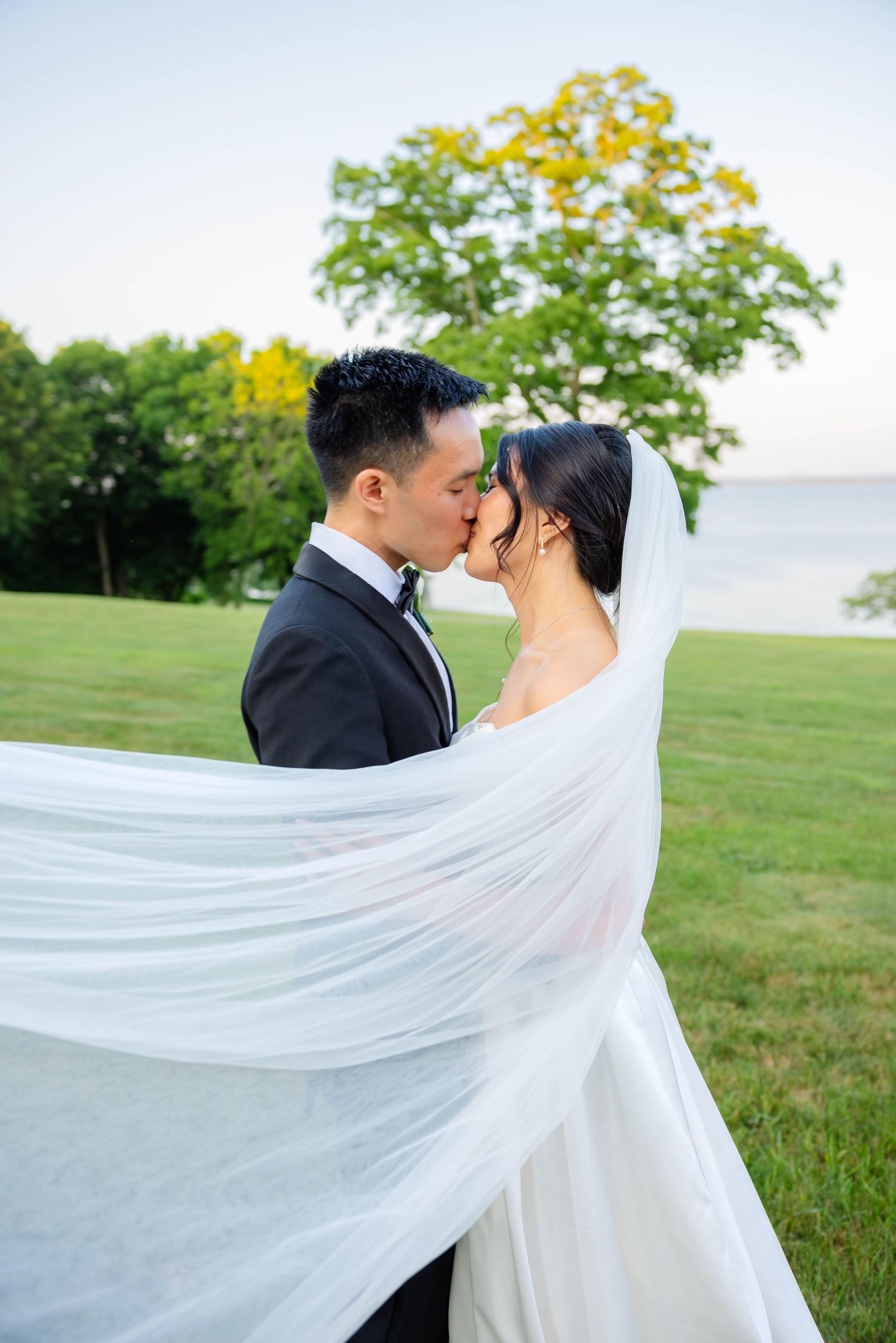 Bride and groom share a kiss outdoors as flowing white veil billows around them near waterfront.