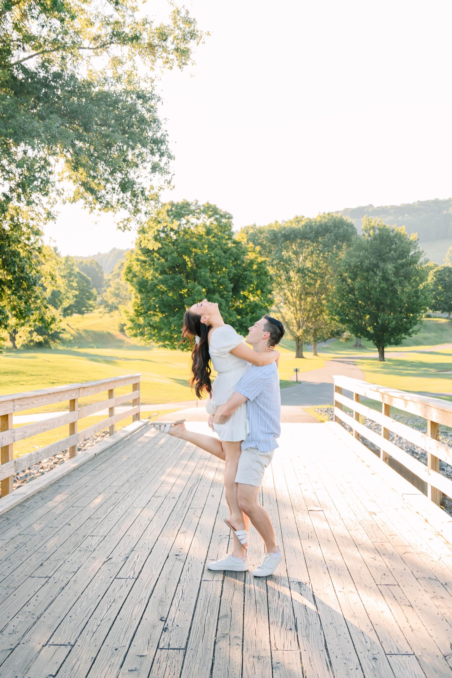 Man lifting woman on a wooden bridge surrounded by golden sunlight and green trees.