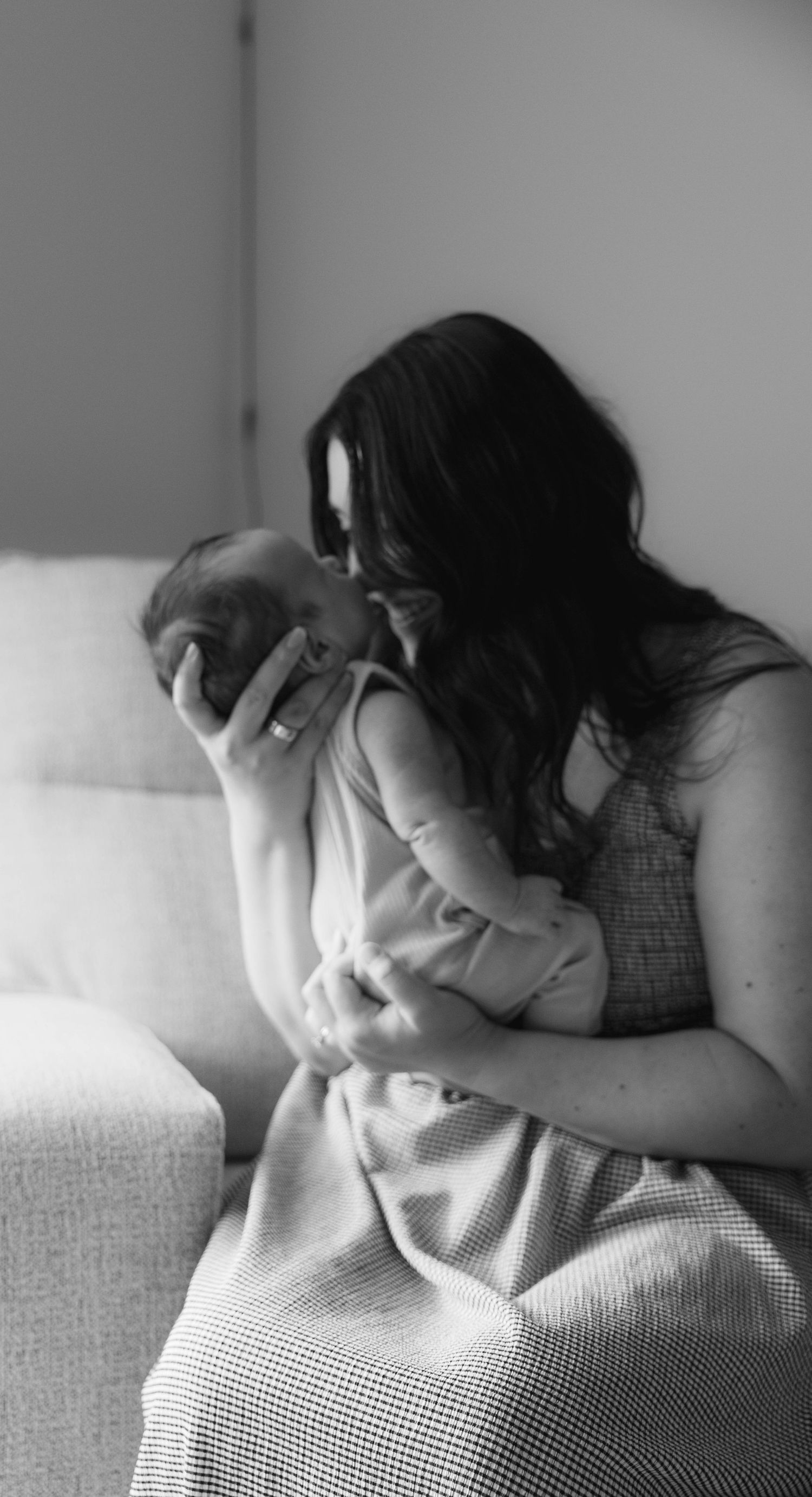 Black and white photo of a dark-haired mother tenderly kissing her newborn baby while sitting on a sofa.