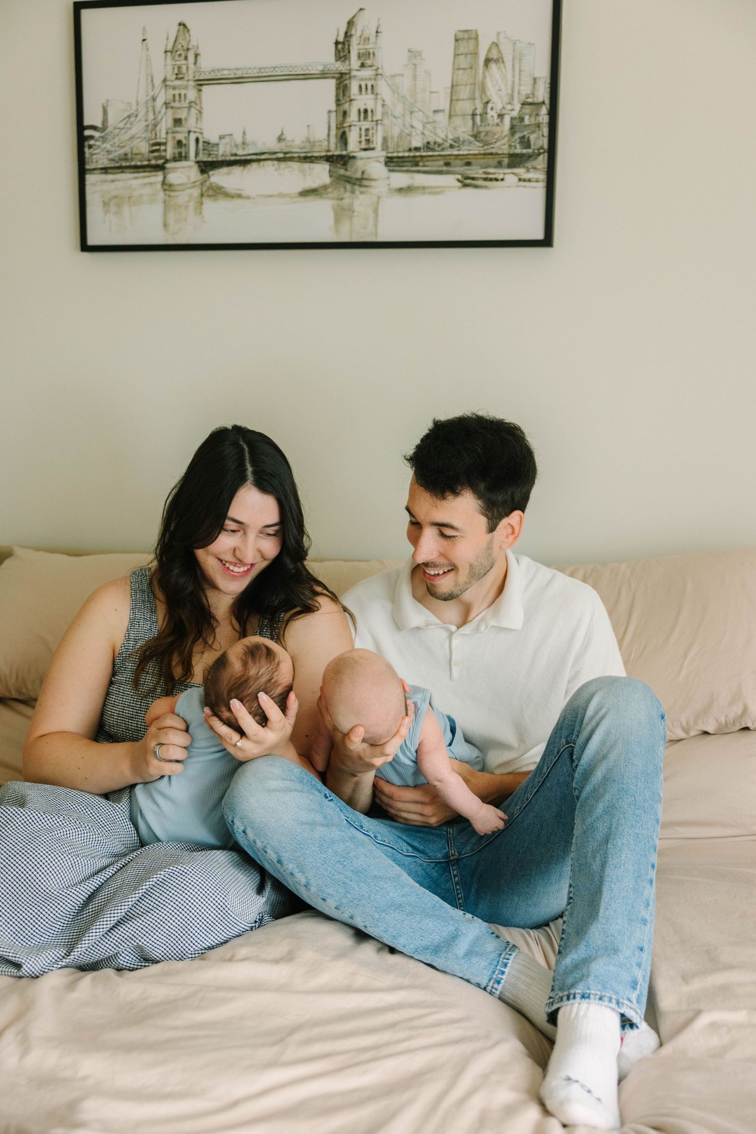 Loving young couple sits on a bed admiring their newborn baby with a London Bridge artwork above.