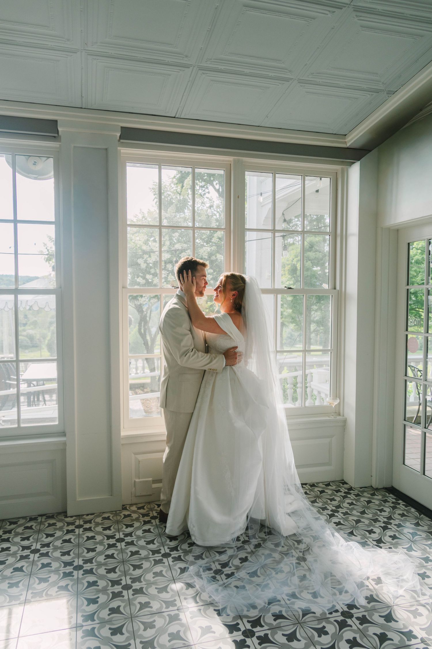 Bride and groom embracing near large windows in elegant room with patterned tile floor and white wedding dress train.