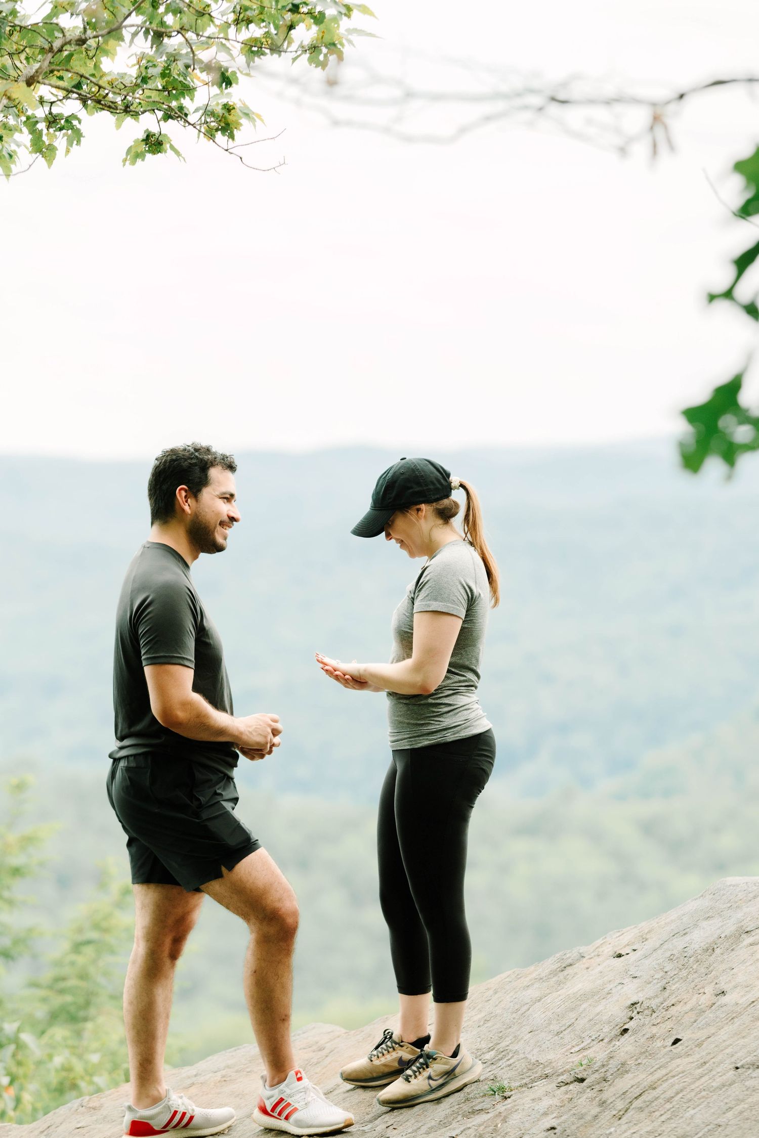 Couple standing on a mountain overlook as woman examines an engagement ring with man smiling nearby.