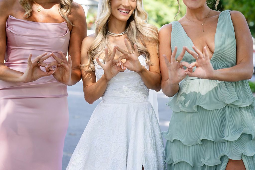 Bride in white dress flanked by two bridesmaids in pink and sage green making heart hand gestures.