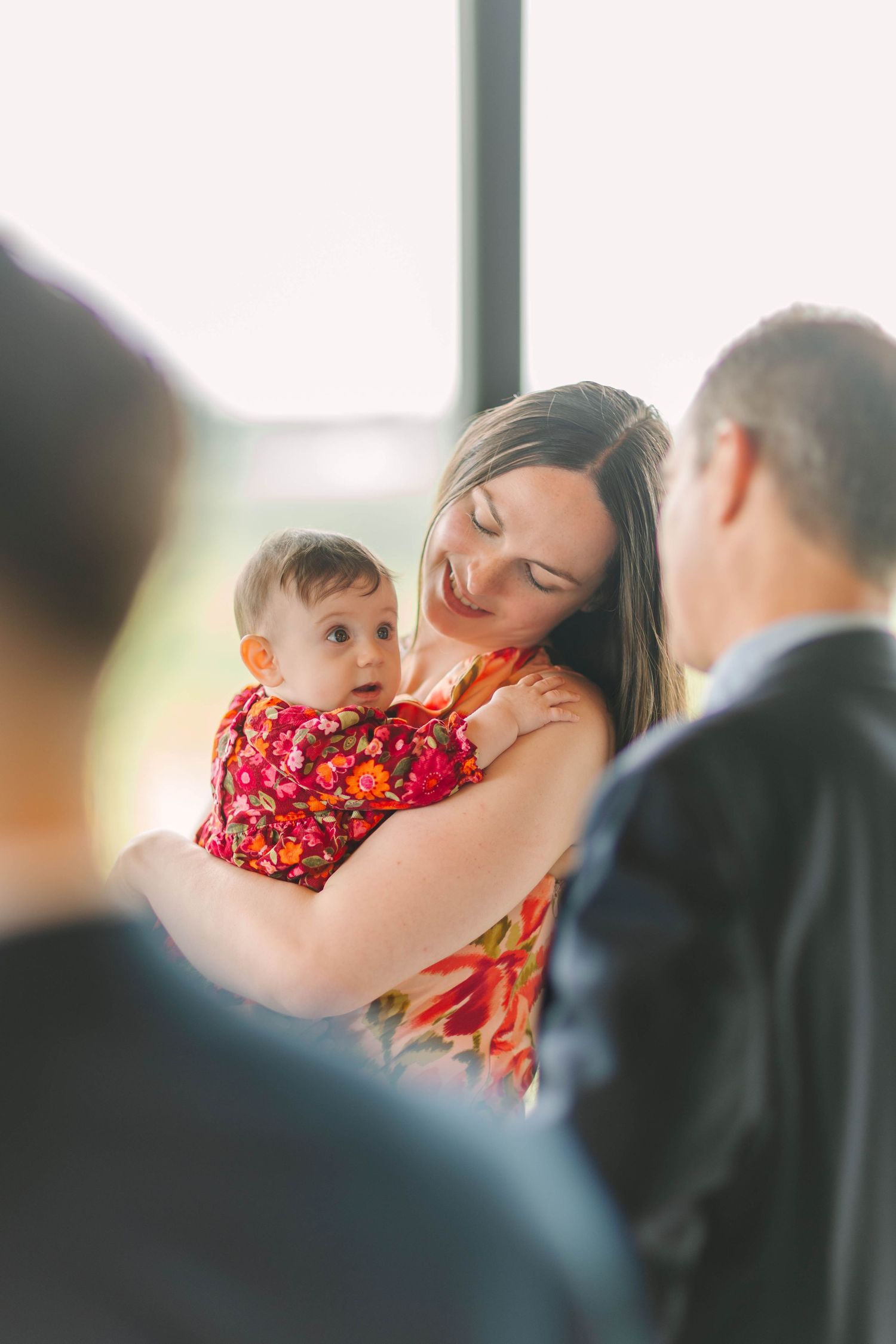 Smiling mother in floral dress tenderly holds her baby girl in red floral outfit at a wedding reception.