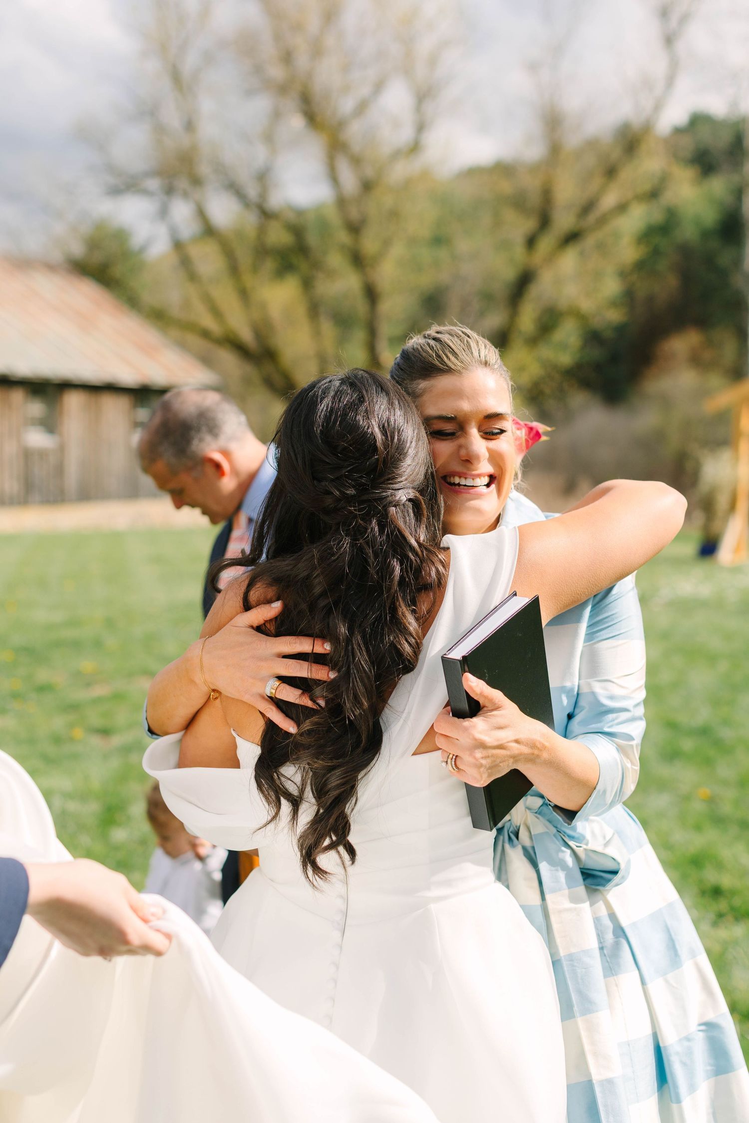 Bride in white gown sharing joyful hug with smiling guest in blue dress holding black book at outdoor wedding.
