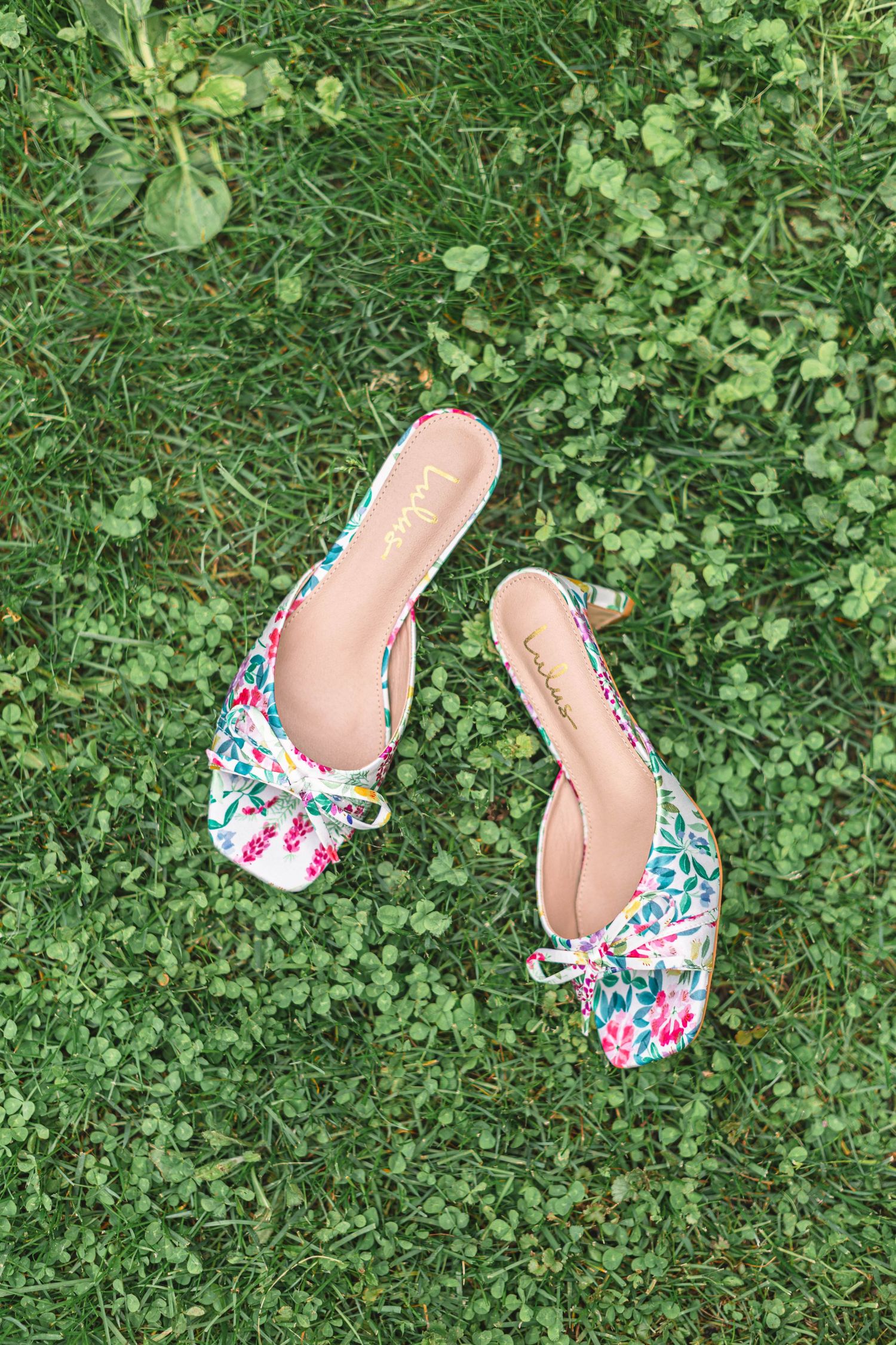 Floral embroidered mule wedding shoes with pink and green design laid on green grass.